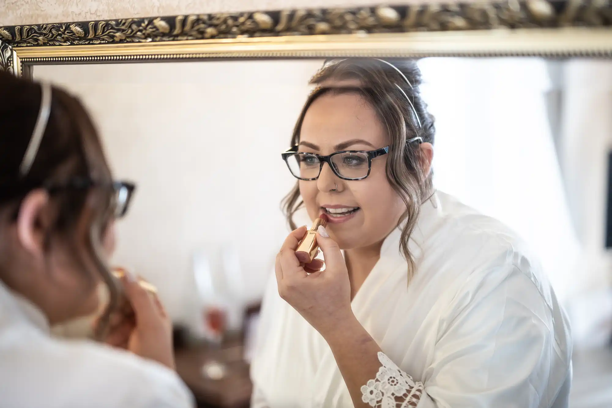 A woman in a white dressing gown and glasses applies lipstick while looking into a large ornate mirror. Her hair is up and she is smiling slightly. The scene suggests preparation for a special occasion, possibly a wedding or formal event.