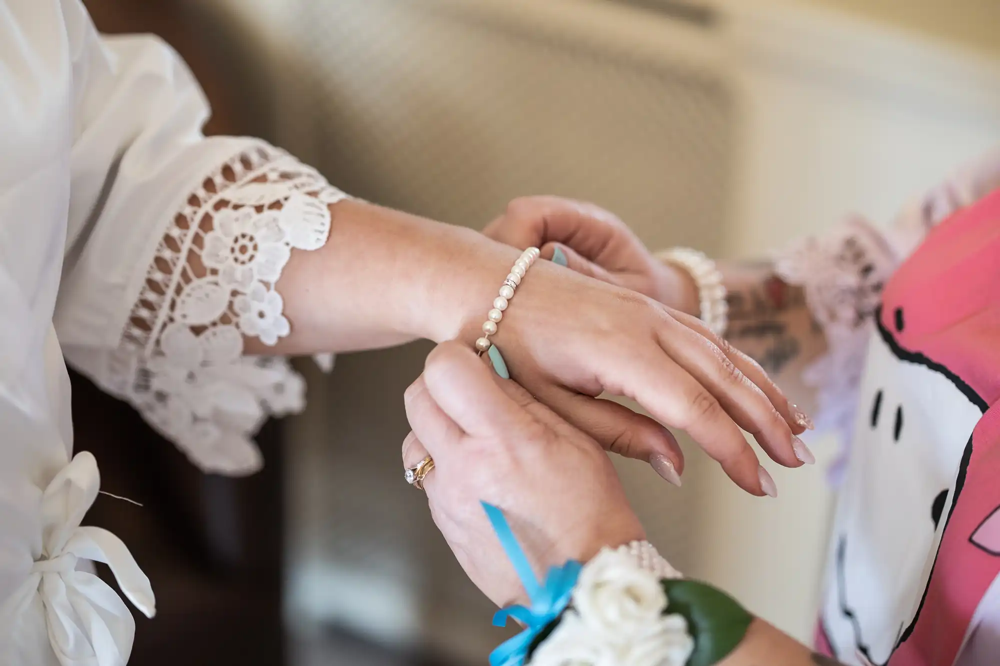 Close-up of one person fastening a pearl bracelet on another’s wrist. Both wear floral lace cuffs, and one has a white rose corsage. The scene suggests a special occasion, possibly a wedding, with gentle natural lighting and soft focus on the hands.