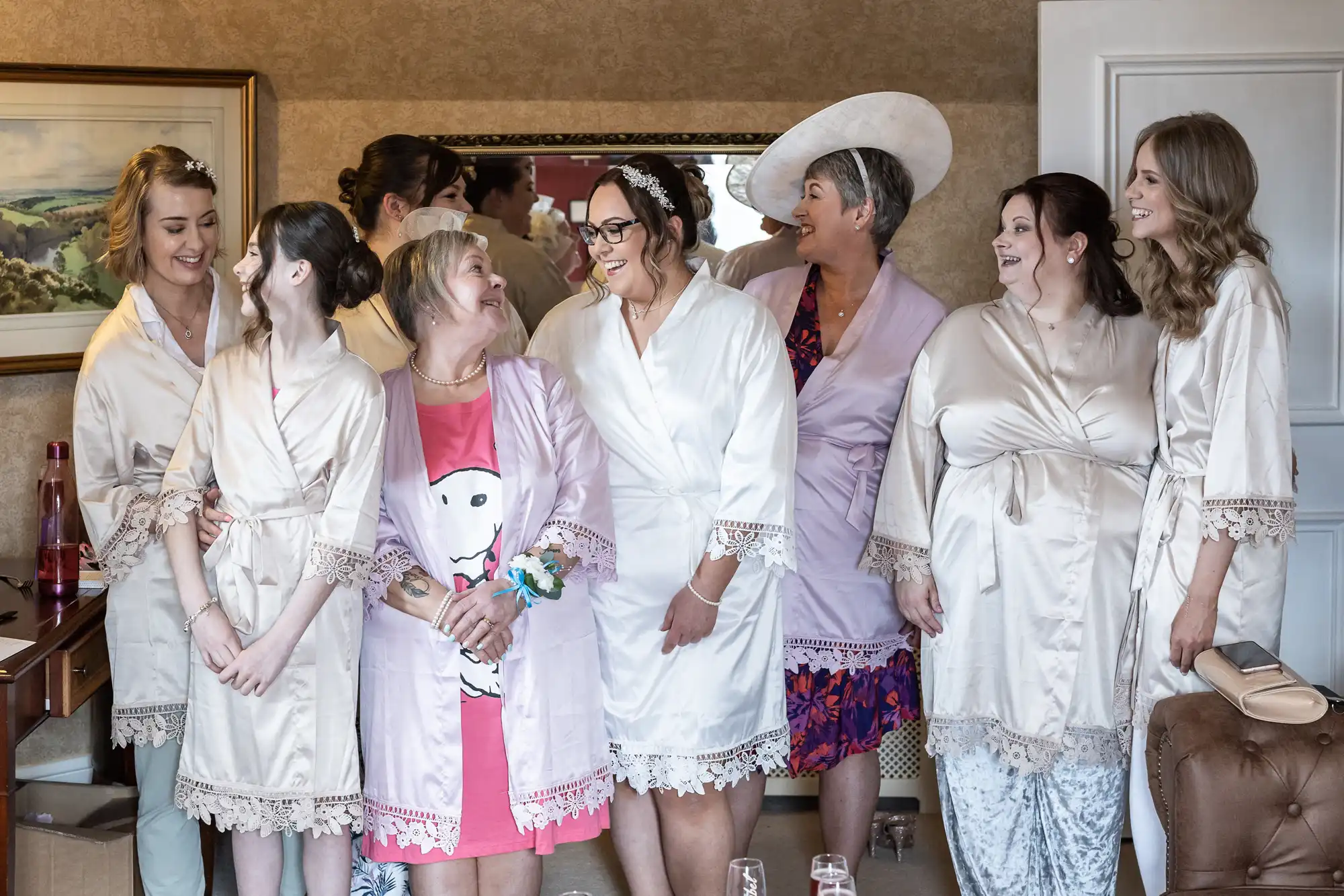 Eight women stand in a room, smiling and talking. Most wear cream or light pink dressing gowns with lace trim, while one at the front wears a bright pink dress with a white cartoon dog. One woman wears a large white hat. The scene is warm and joyful, suggesting wedding preparations.