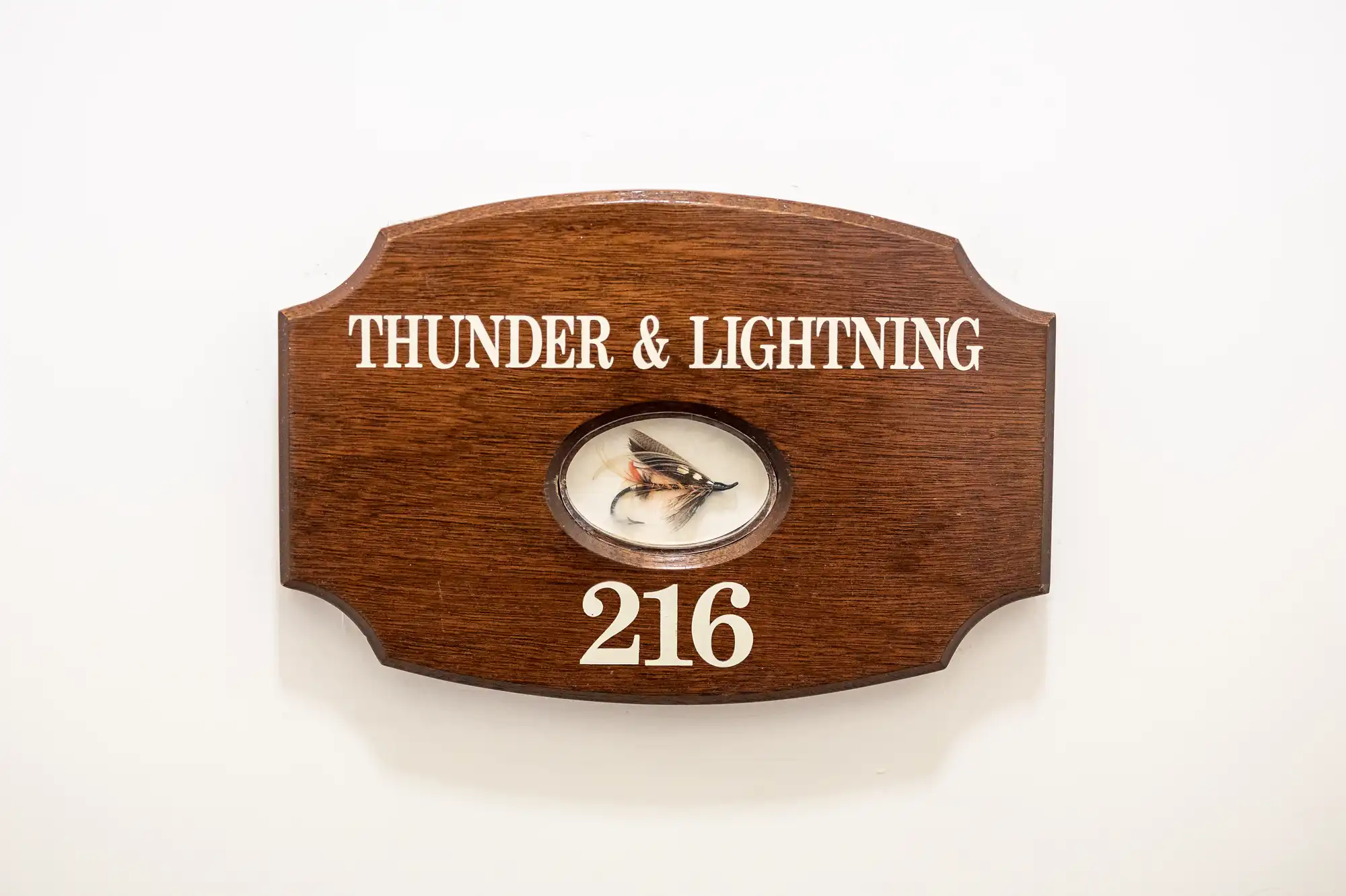 A wooden plaque with white text reads "THUNDER & LIGHTNING" above an oval inset displaying a fishing fly. Below the inset, the number "216" is printed. The plaque is mounted on a plain white wall.