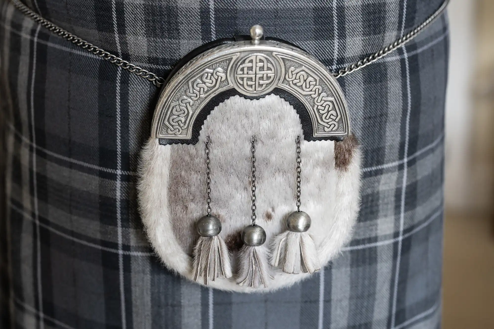 A traditional Scottish sporran with Celtic knotwork on a silver top, fur body, and three tassels hangs from a chain over a grey and white checked kilt.