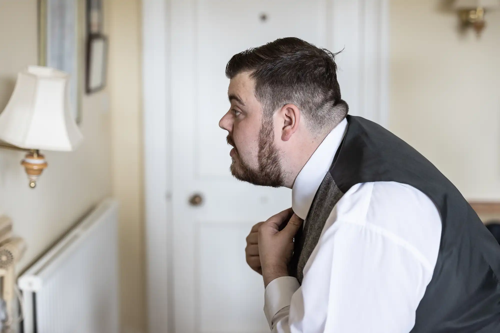 A man with short dark hair and beard, dressed in a white shirt and dark waistcoat, stands indoors adjusting his tie in front of a mirror. The room is softly lit, with a lamp and radiator visible in the background.