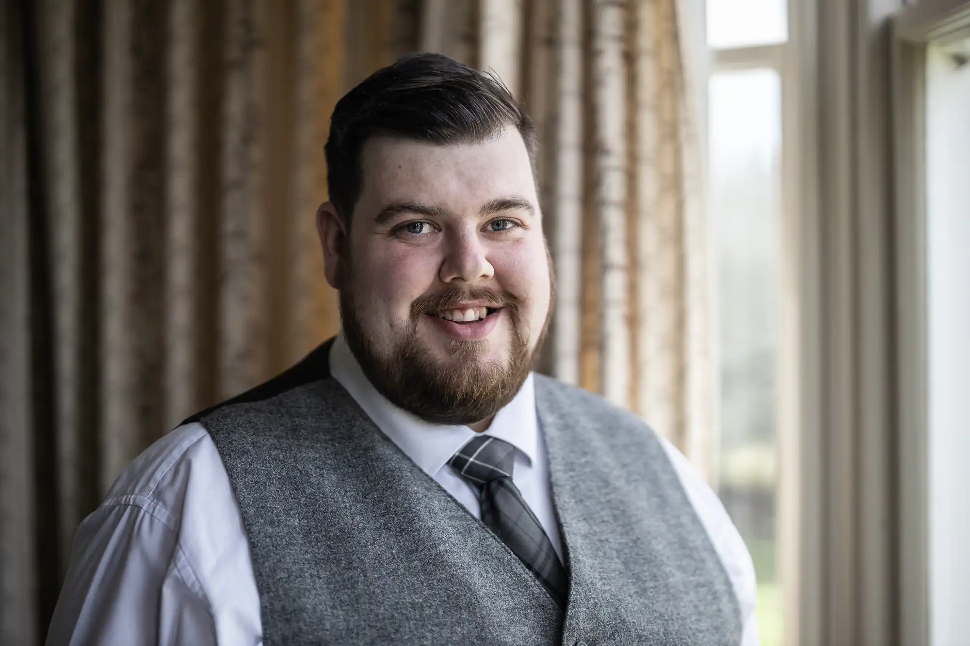 A man with short dark hair and a beard smiles, wearing a white shirt, grey waistcoat, and striped tie. He stands indoors near a window with beige patterned curtains and soft natural light illuminating his face.