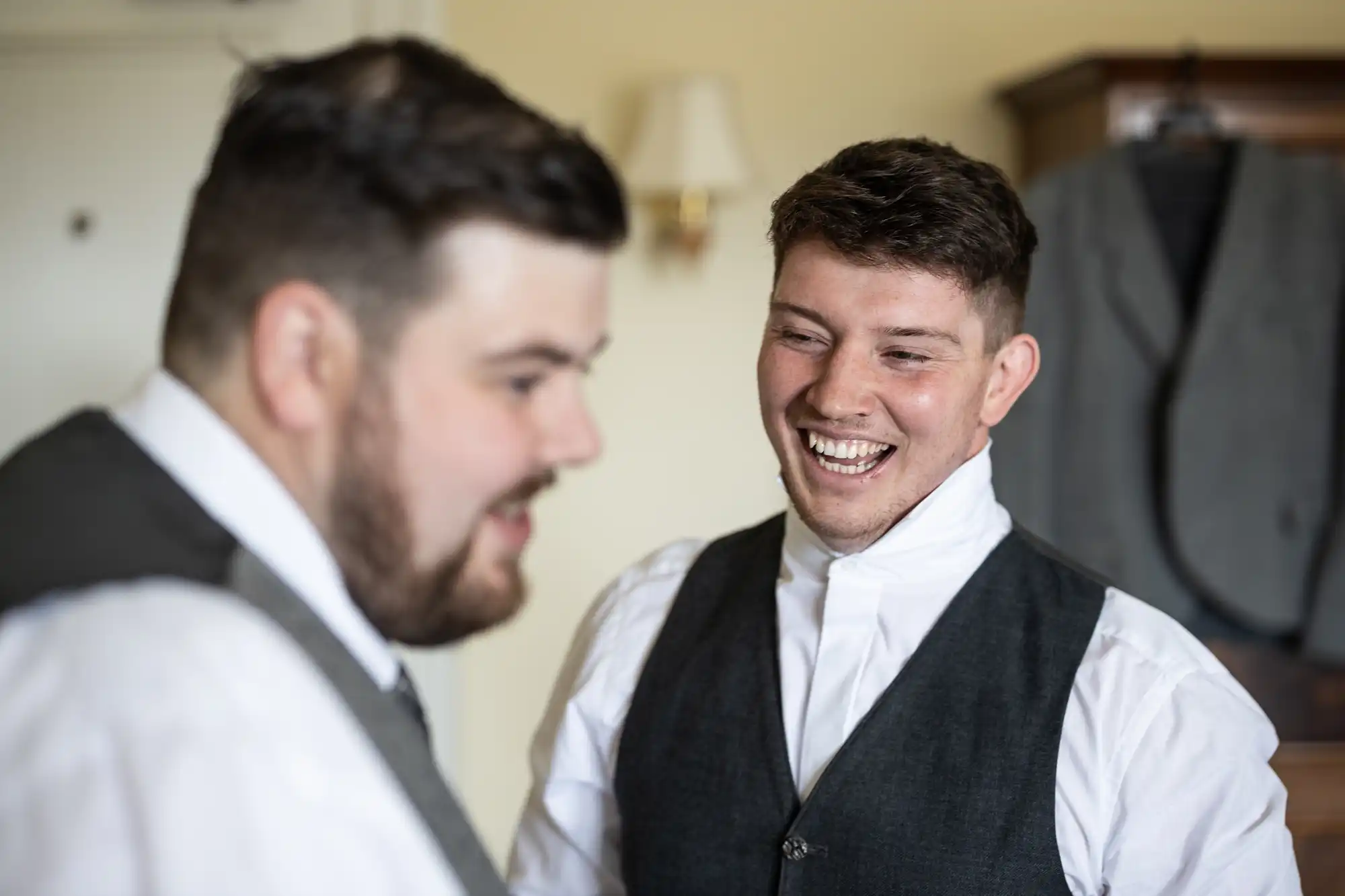 Two men in formal attire, both wearing white shirts and dark waistcoats, smile and laugh together indoors. One man is in sharp focus, grinning widely, while the other is blurred in the foreground. A suit jacket hangs in the background.