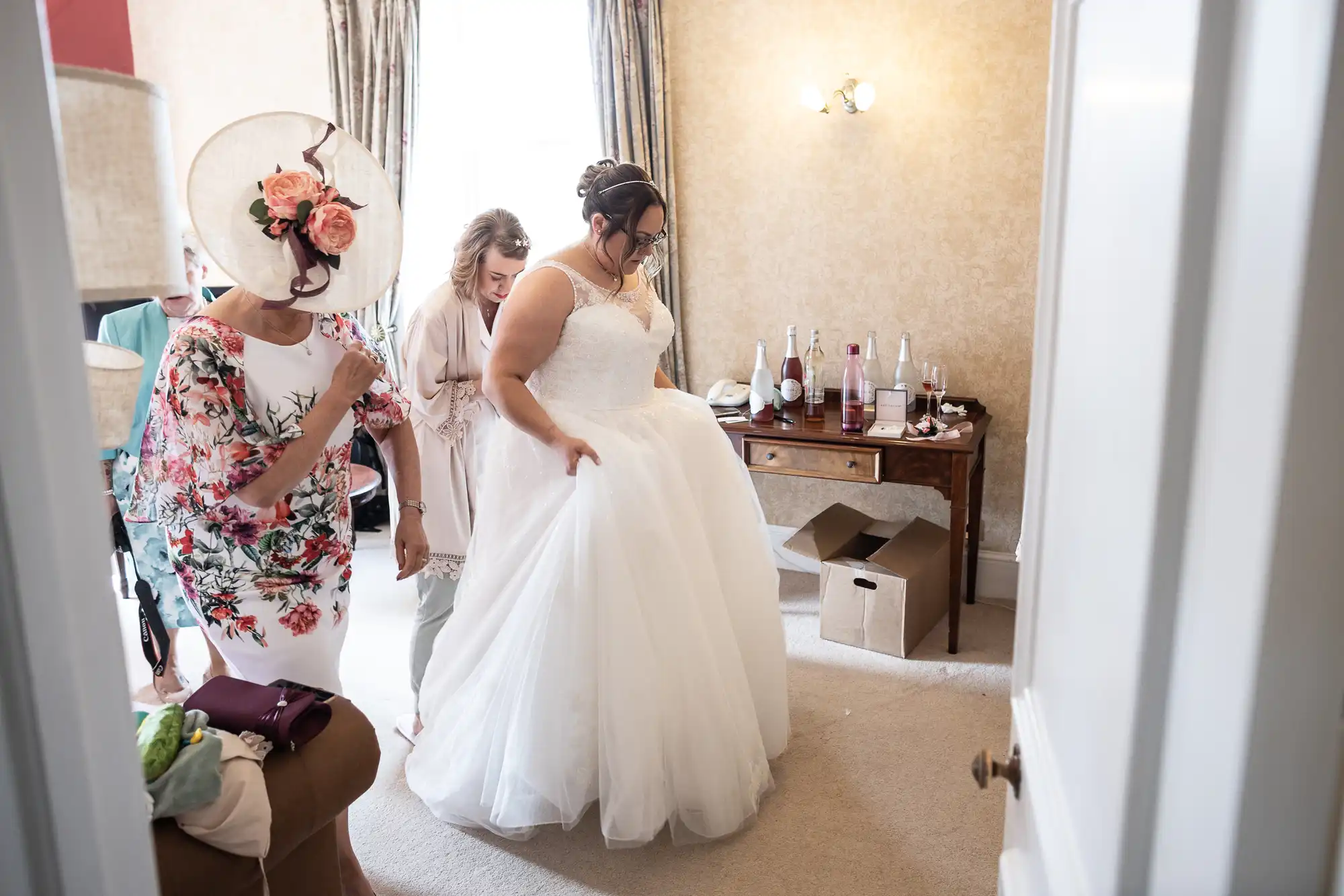 A bride in a white gown stands in a softly lit room as two women help adjust her dress. One woman wears a floral outfit with a wide hat; the other is in pastel attire. Behind them is a desk with bottles and glasses, and light pours in from tall windows.
