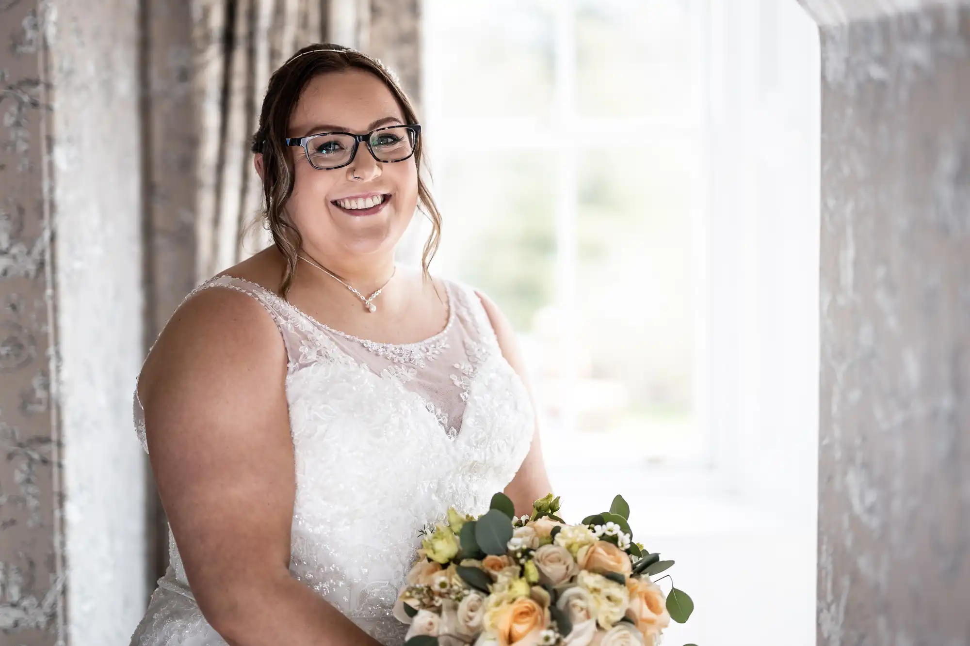 A smiling bride with glasses and brown hair, wearing a white lace wedding dress, holds a bouquet of yellow and white roses with greenery. She is seated indoors near a window with light curtains and soft natural light illuminating her face.
