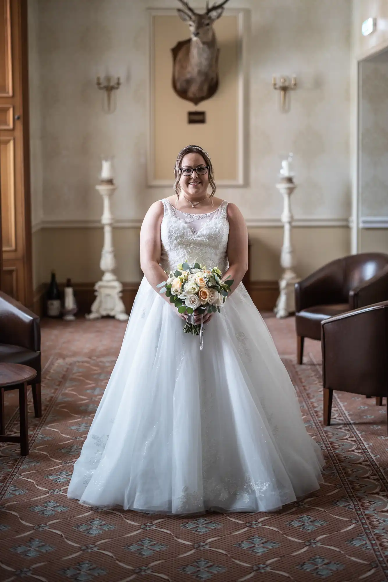 A bride in a white, sleeveless wedding gown with lace details holds a pastel bouquet and smiles at the camera. She stands in an elegant room with brown chairs, tall candlesticks, and a mounted stag’s head on the cream-coloured wall behind her.