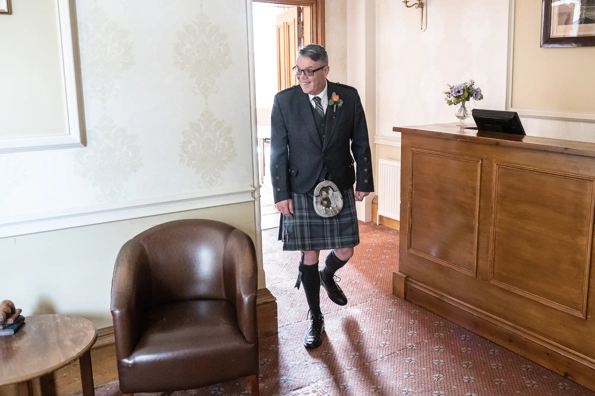 A man wearing glasses and a traditional Scottish kilt outfit smiles as he walks indoors near a wooden reception desk. The room has light wallpaper, carpeted floor, a brown leather chair, and a small flower arrangement on the counter.
