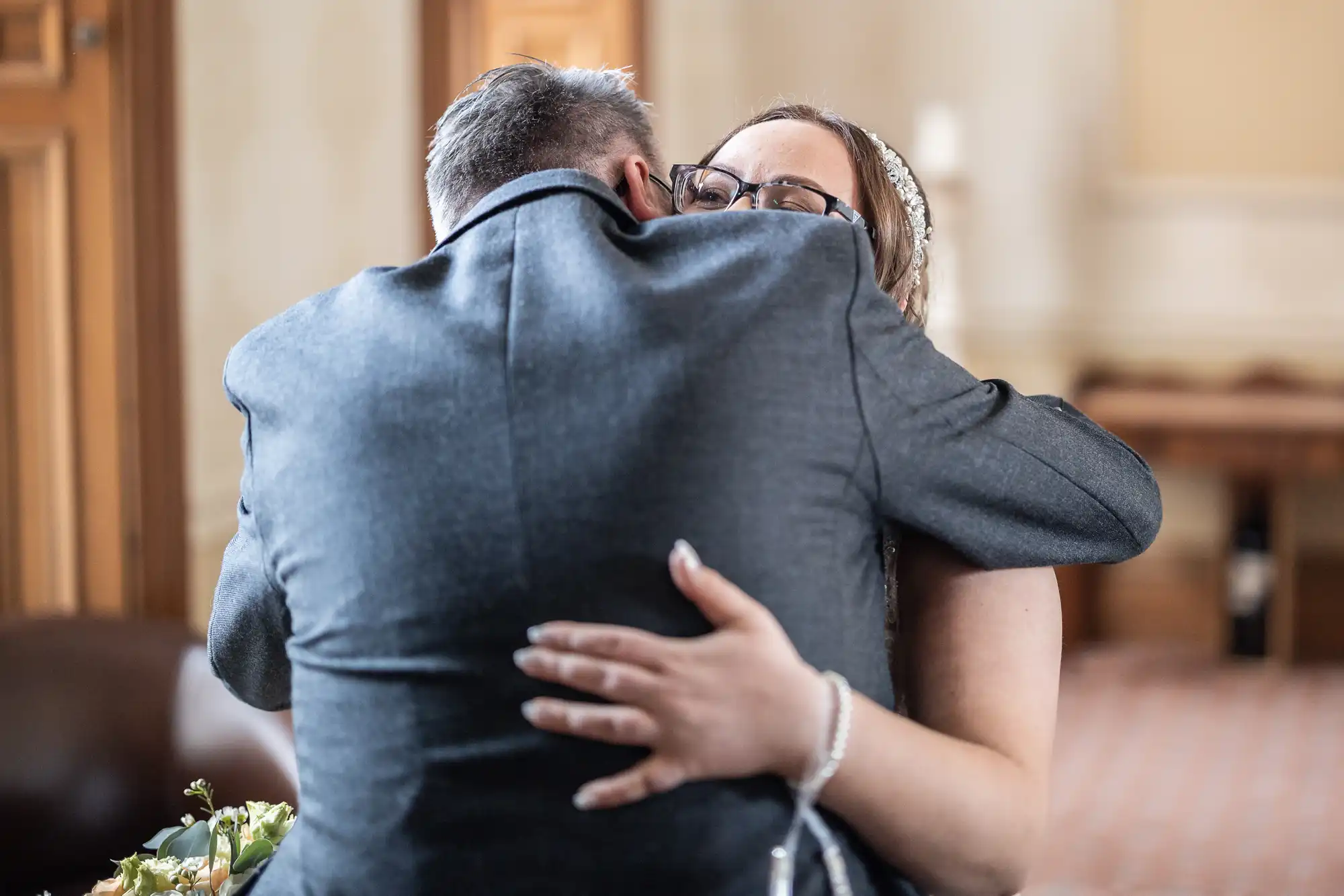 A man in a grey suit hugs a woman with long brown hair, glasses, and a jewelled headband. The woman smiles and closes her eyes, one hand visible on the man's back, showing a bracelet and manicured nails. The background is softly blurred.