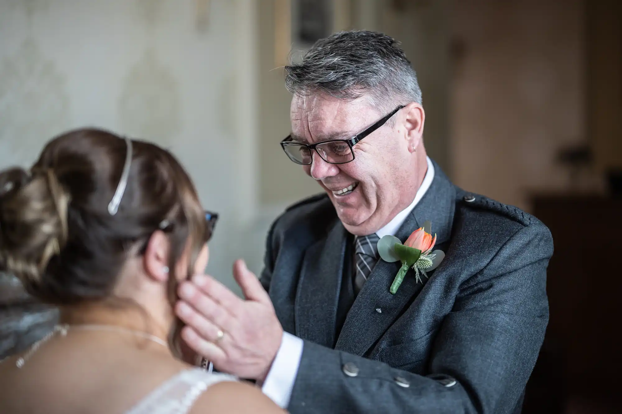 An older man in a grey suit and glasses smiles warmly, touching the face of a woman in a wedding dress with a jewelled hairpiece. The man wears a buttonhole with a peach tulip. They share an emotional moment indoors.