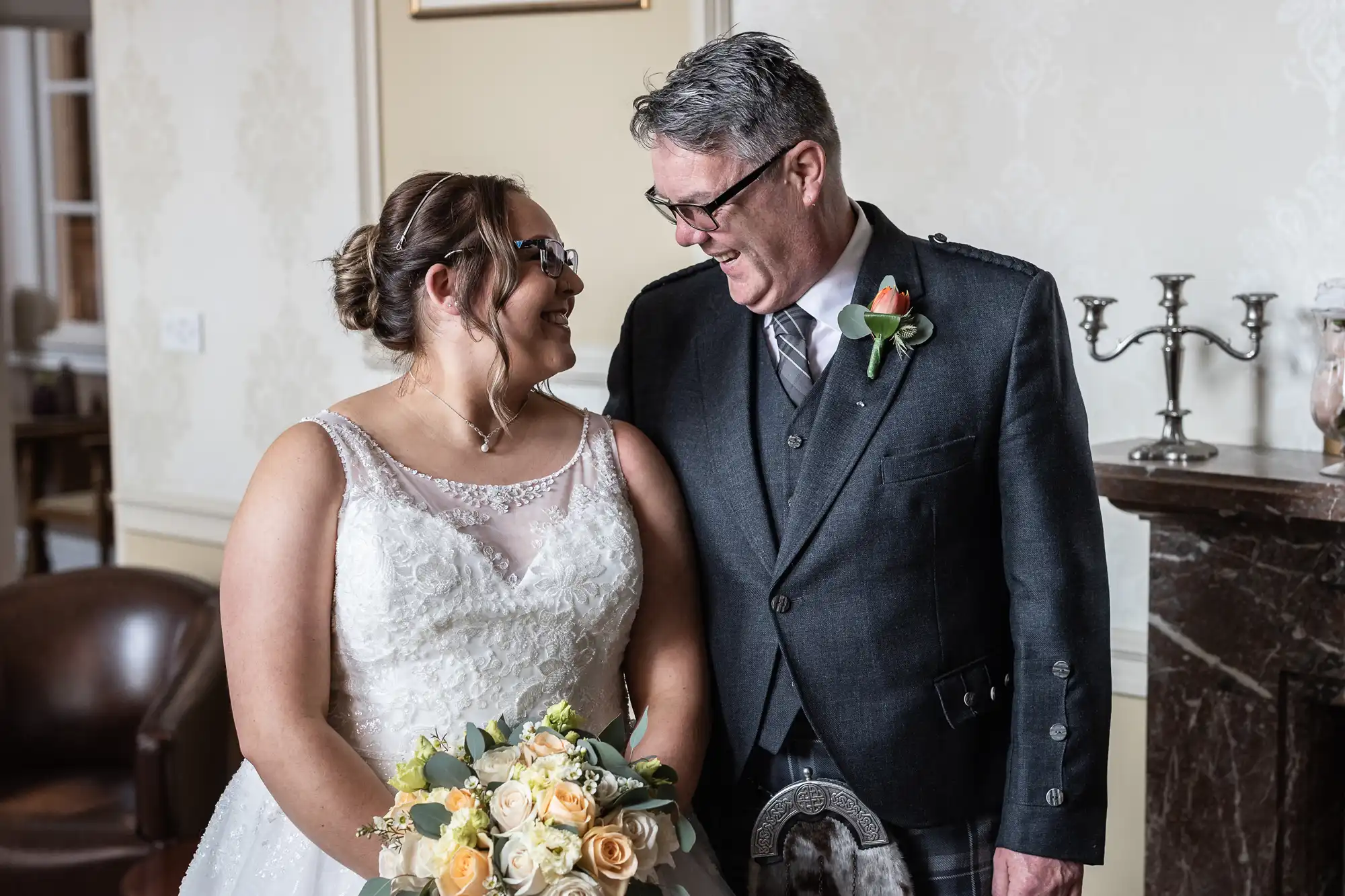 A bride in a white gown and glasses smiles warmly at an older man in a dark suit and buttonhole. The bride holds a bouquet of yellow and white roses. They stand indoors, appearing joyful, facing each other beside a brown leather chair and a fireplace.