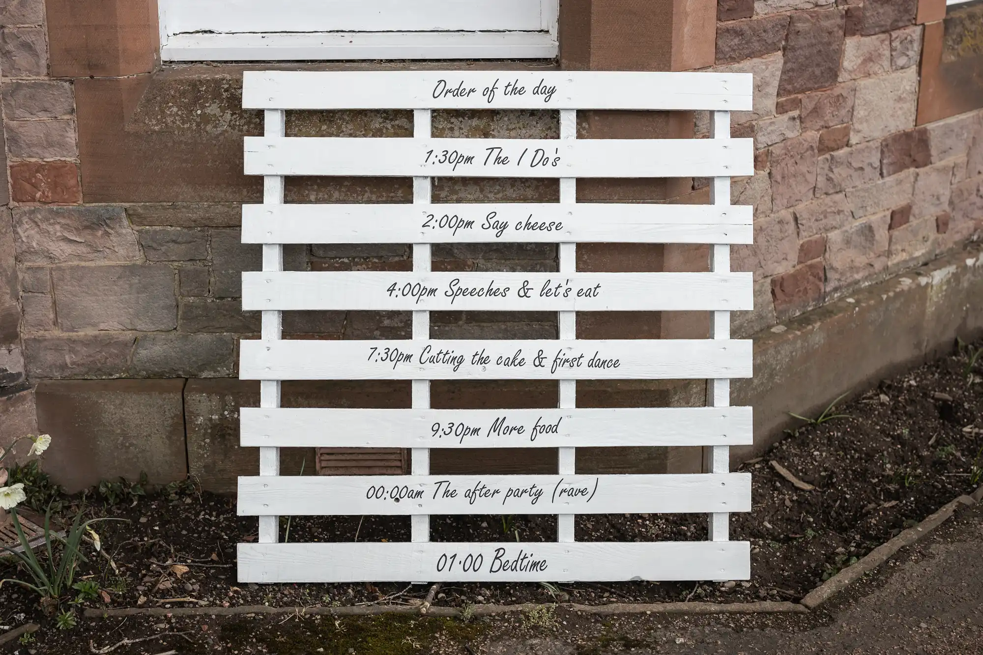 A rustic wedding schedule is painted in white on a wooden pallet, leaning against a stone wall. The schedule includes times and events such as: "Order of the day, Say cheese, Speeches & let’s eat, Cutting the cake & first dance, More food, After party, Bedtime.