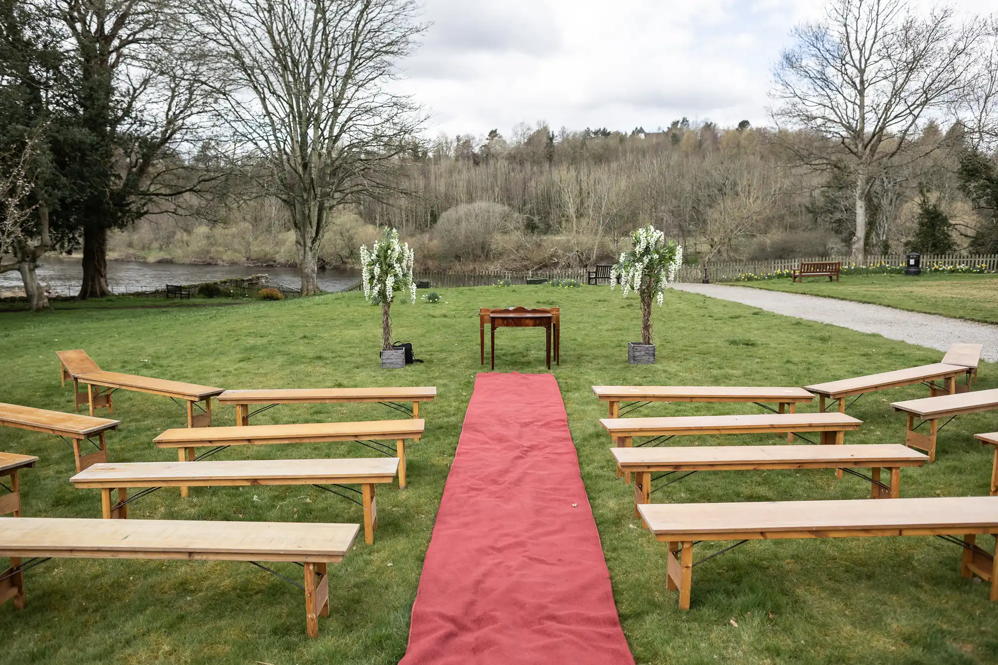 Outdoor wedding setup with wooden benches on grass, a red carpet aisle leading to a small wooden table between two flower-adorned arches, and trees with bare branches in the background under a cloudy sky.