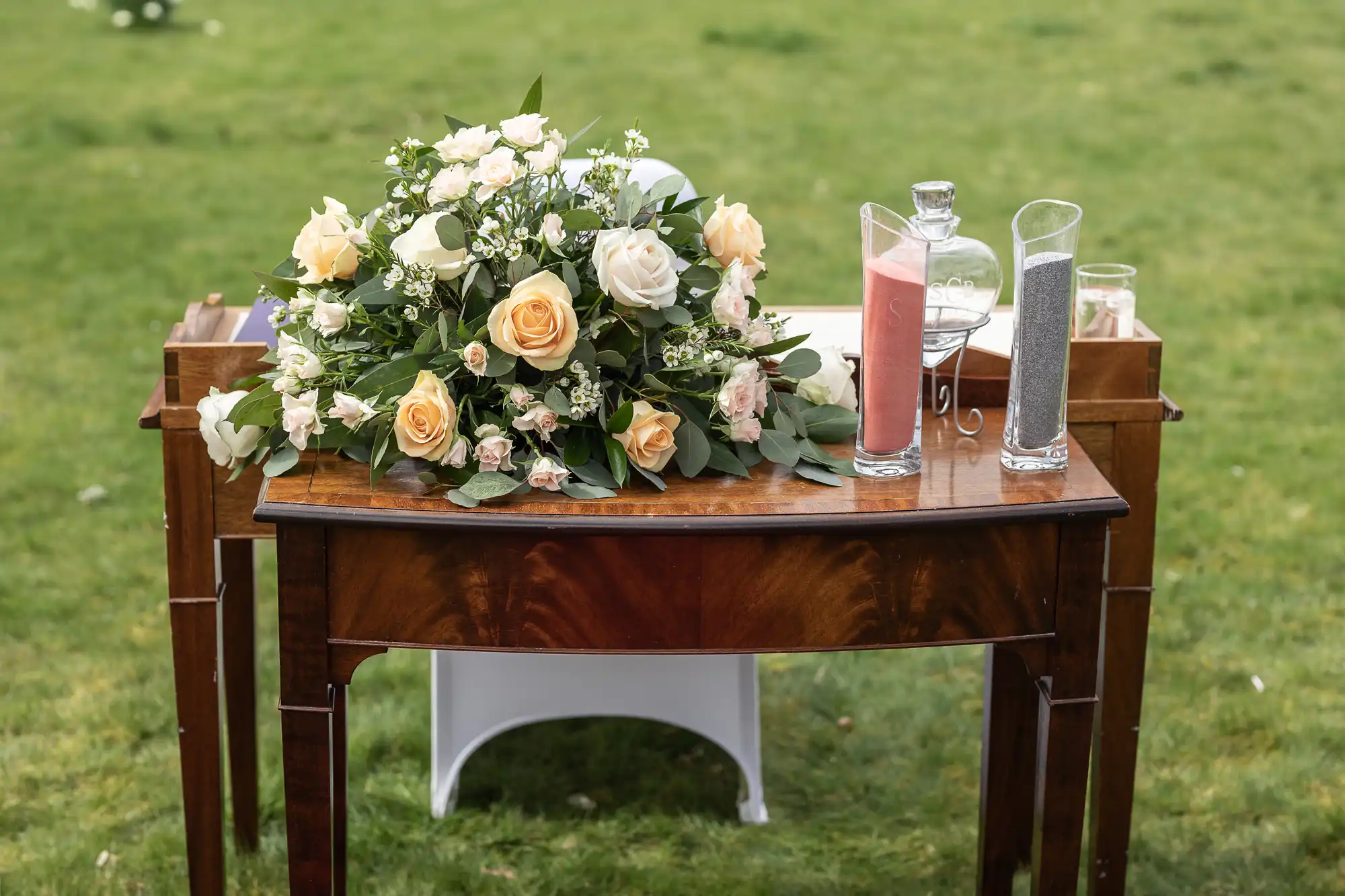 A wooden table is decorated with a bouquet of yellow and white roses with greenery. Beside the flowers are two glass vases filled with pink and grey sand, and a glass decanter, all set outdoors on green grass.