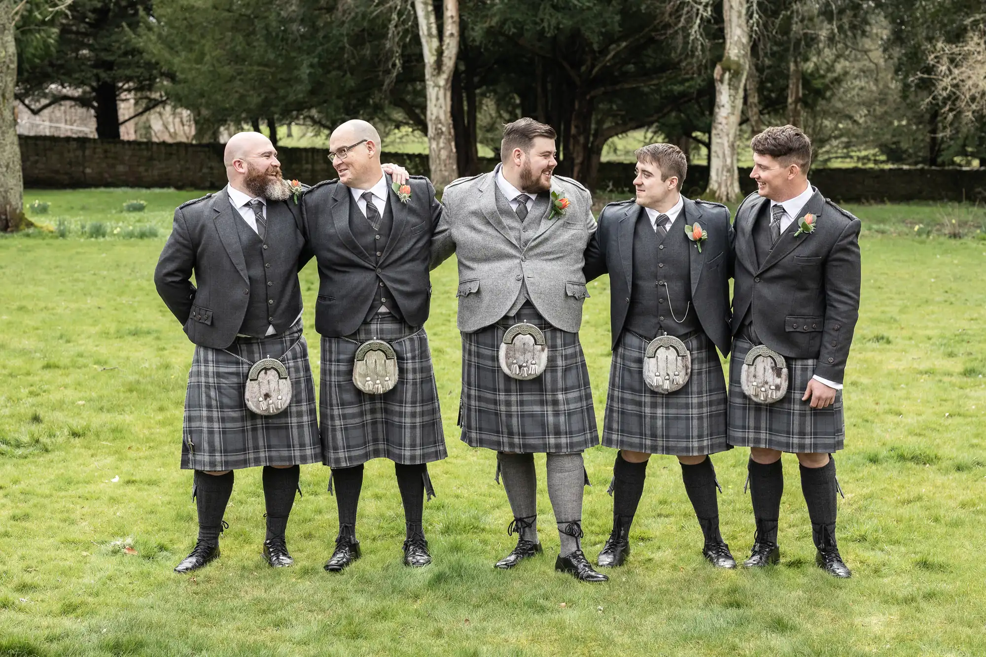 Five men stand on grass, smiling at each other. They wear matching dark tartan kilts, jackets, sporrans, and long socks. The man in the centre wears a lighter grey jacket. There are trees and a stone wall in the background, suggesting an outdoor event.