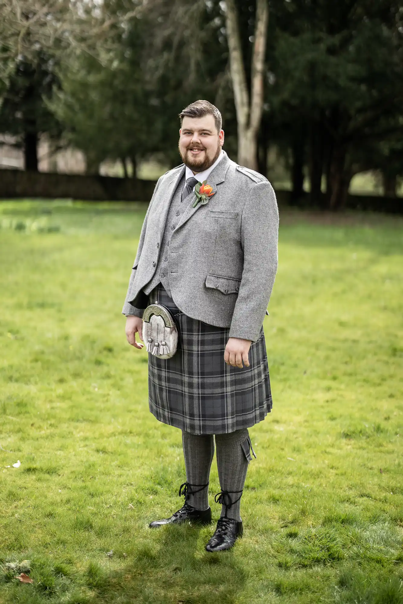 A man stands on green grass, smiling, wearing a grey tweed jacket, matching waistcoat, and a grey tartan kilt with knee-high socks and black shoes. He has a sporran at his waist and an orange rose buttonhole. Trees and a stone wall are in the background.