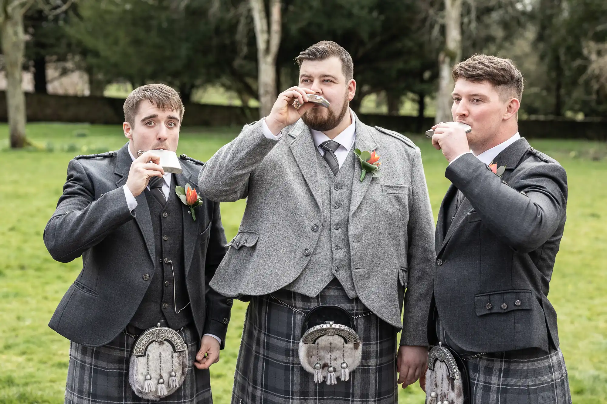 Three men wearing grey jackets, waistcoats, kilts, and sporrans stand outdoors on grass, drinking from small glasses. Each has a flower buttonhole. Trees and a stone wall are blurred in the background. All look towards the camera.