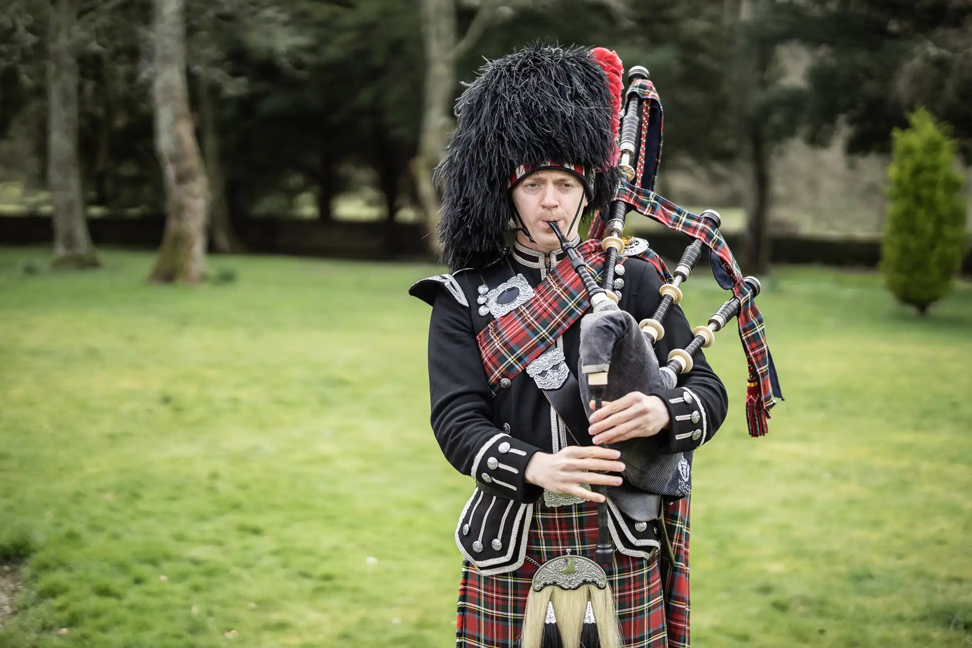A man in traditional Scottish attire, including a feathered hat and tartan kilt, stands on grass playing the bagpipes. Trees and shrubs are in the background. He looks forward while holding and blowing into the bagpipes, with tartan fabric draped over his shoulder.