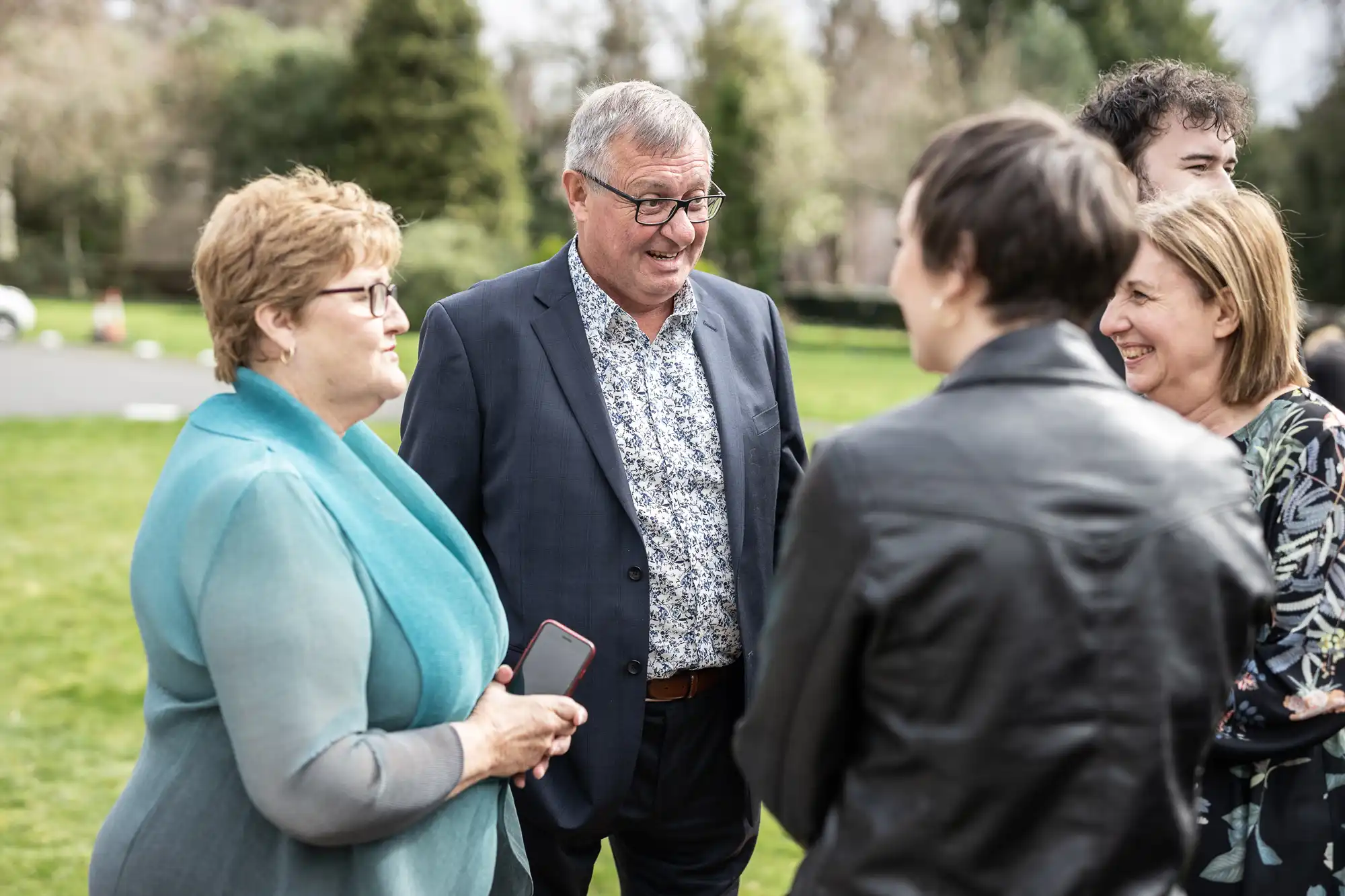 A group of adults stand outside on grass, smiling and talking. One man in a patterned shirt and suit jacket is centred, surrounded by four women, some holding mobiles. Trees and a blurred building are in the background. The mood is friendly and relaxed.
