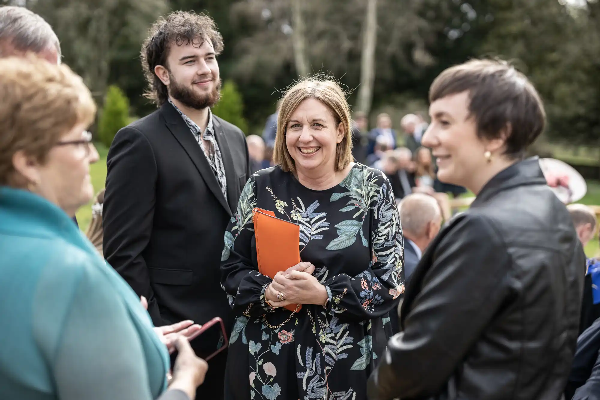 Four adults stand outdoors in conversation. A woman in a black floral dress smiles, holding an orange folder. A young man in a suit stands beside her, smiling. Another woman in a black jacket and a woman in teal are also engaged in the group discussion.