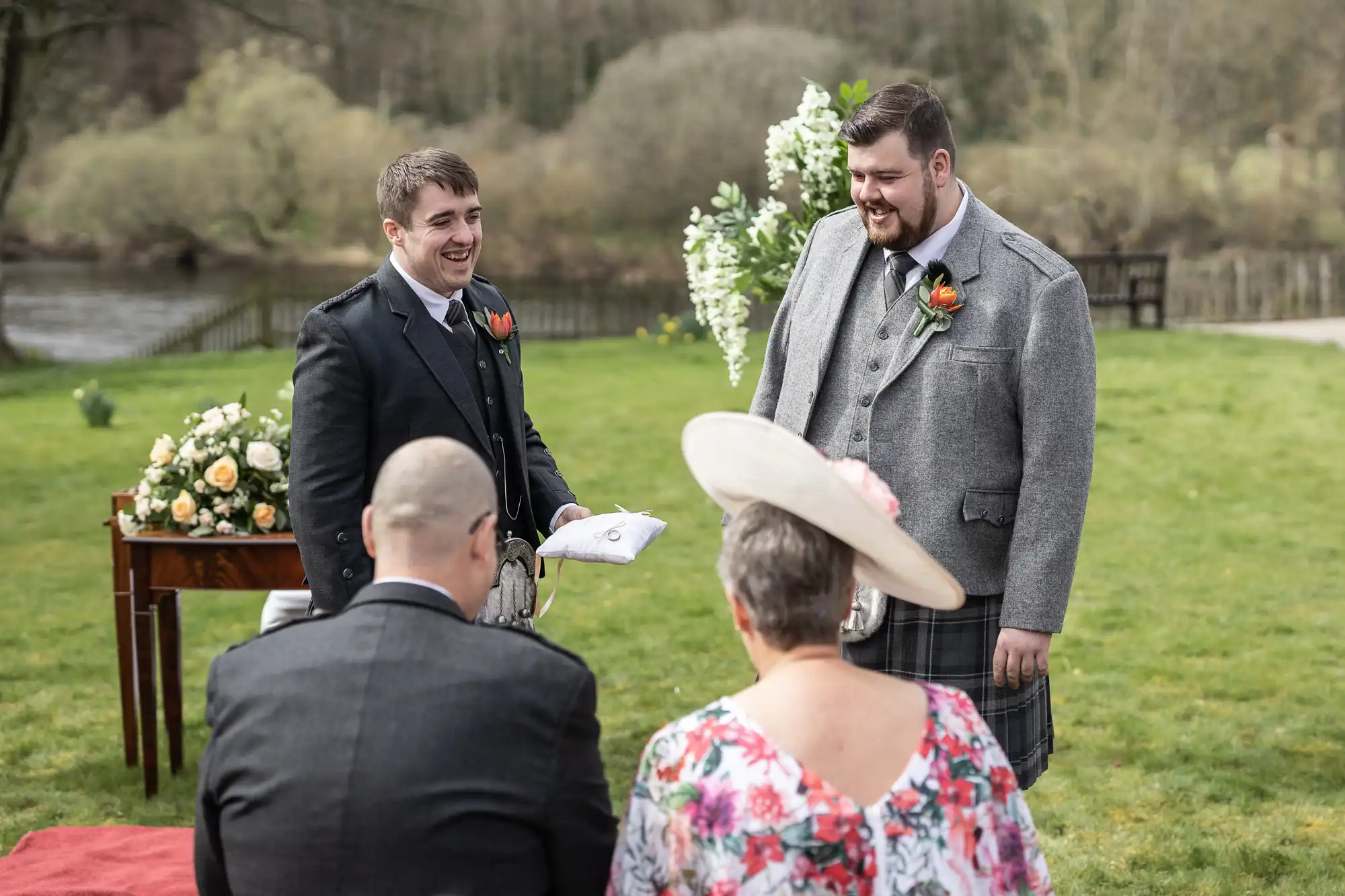 Two men in kilts and jackets stand smiling at each other outdoors, one holding a cloth. They face two seated guests, a man in a suit and a woman in a floral dress with a wide-brimmed hat. An arch with flowers and a table with an arrangement are in the background.