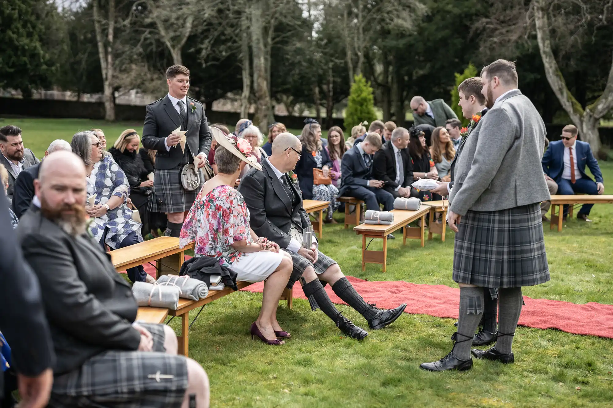 A group of people, many wearing kilts, are seated on wooden benches outdoors for a wedding ceremony. Two men in grey jackets and kilts stand near a red carpet, talking. Trees and greenery are in the background, and the mood is festive and relaxed.