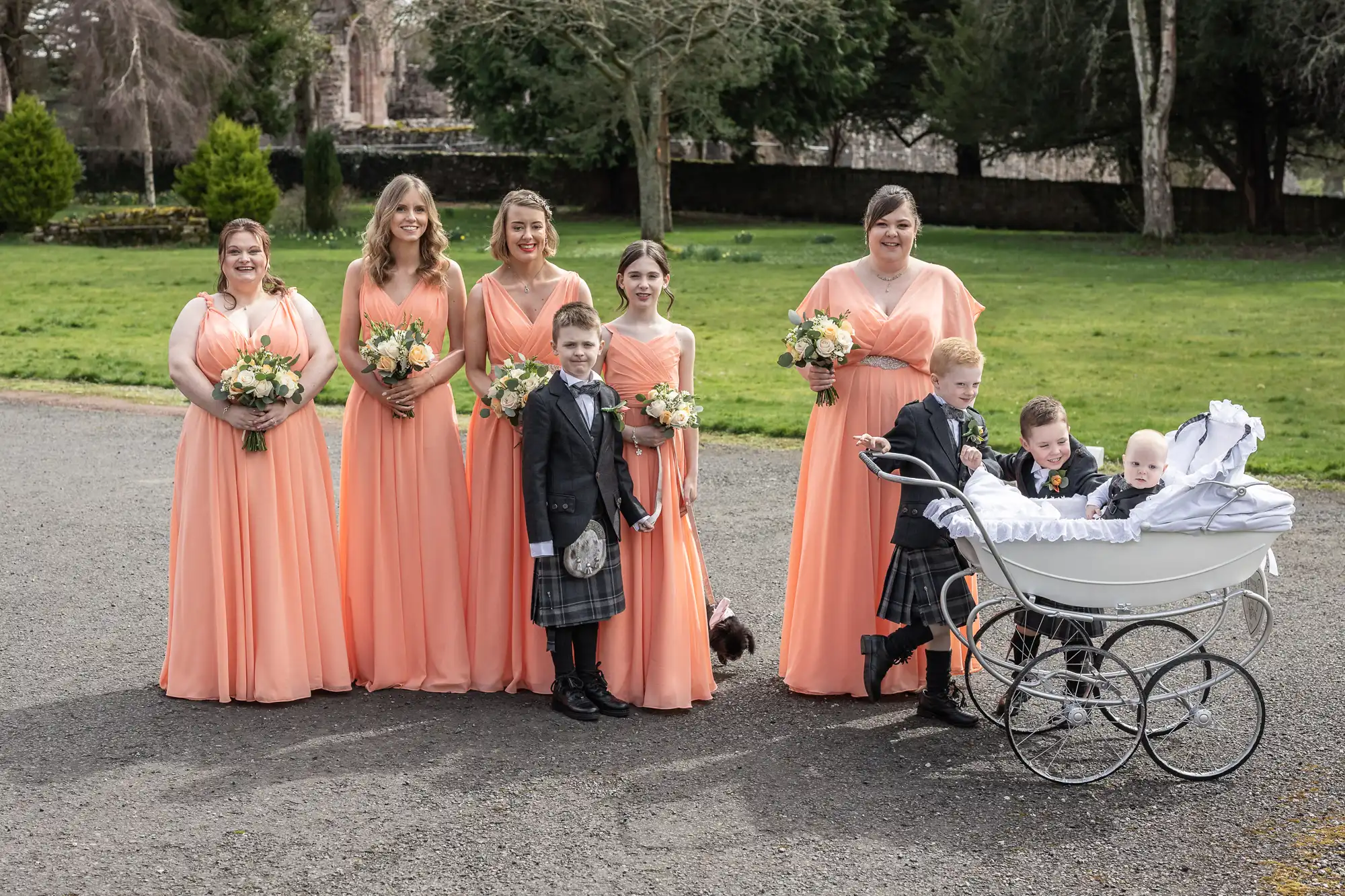 Six bridesmaids in matching peach dresses stand on a path holding bouquets. In front of them, three boys in kilts stand near a vintage white pram with two babies inside. The background shows green grass, trees, and a stone wall.