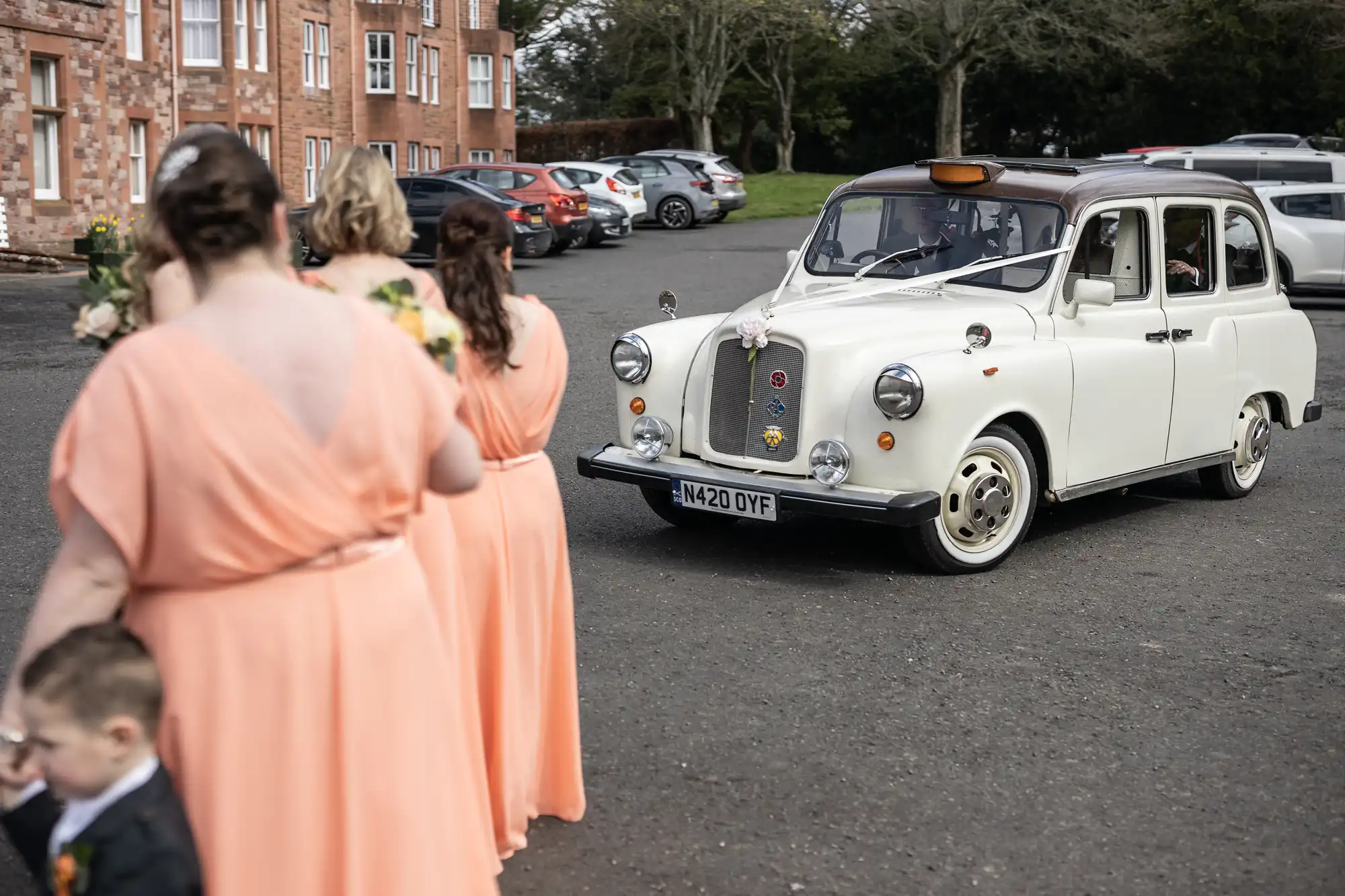 A white vintage taxi decorated with flowers is parked on a paved car park. Three women in peach bridesmaid dresses and a young boy in a suit stand in the foreground with their backs to the camera. Red-brick buildings and parked cars are in the background.