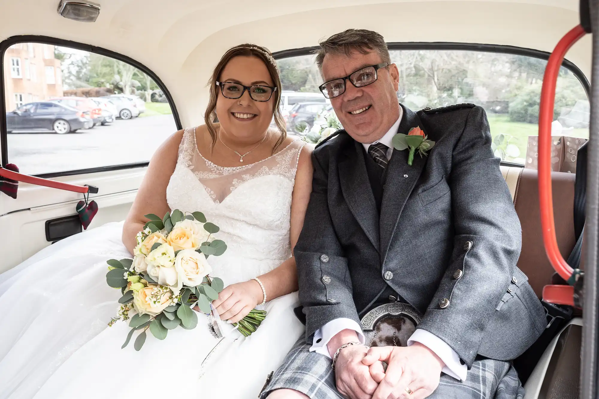 A smiling bride in a white dress holding a bouquet sits beside a man in a grey suit and kilt in the backseat of a classic car. Both look happy and dressed for a wedding, with trees and parked cars visible through the rear window.