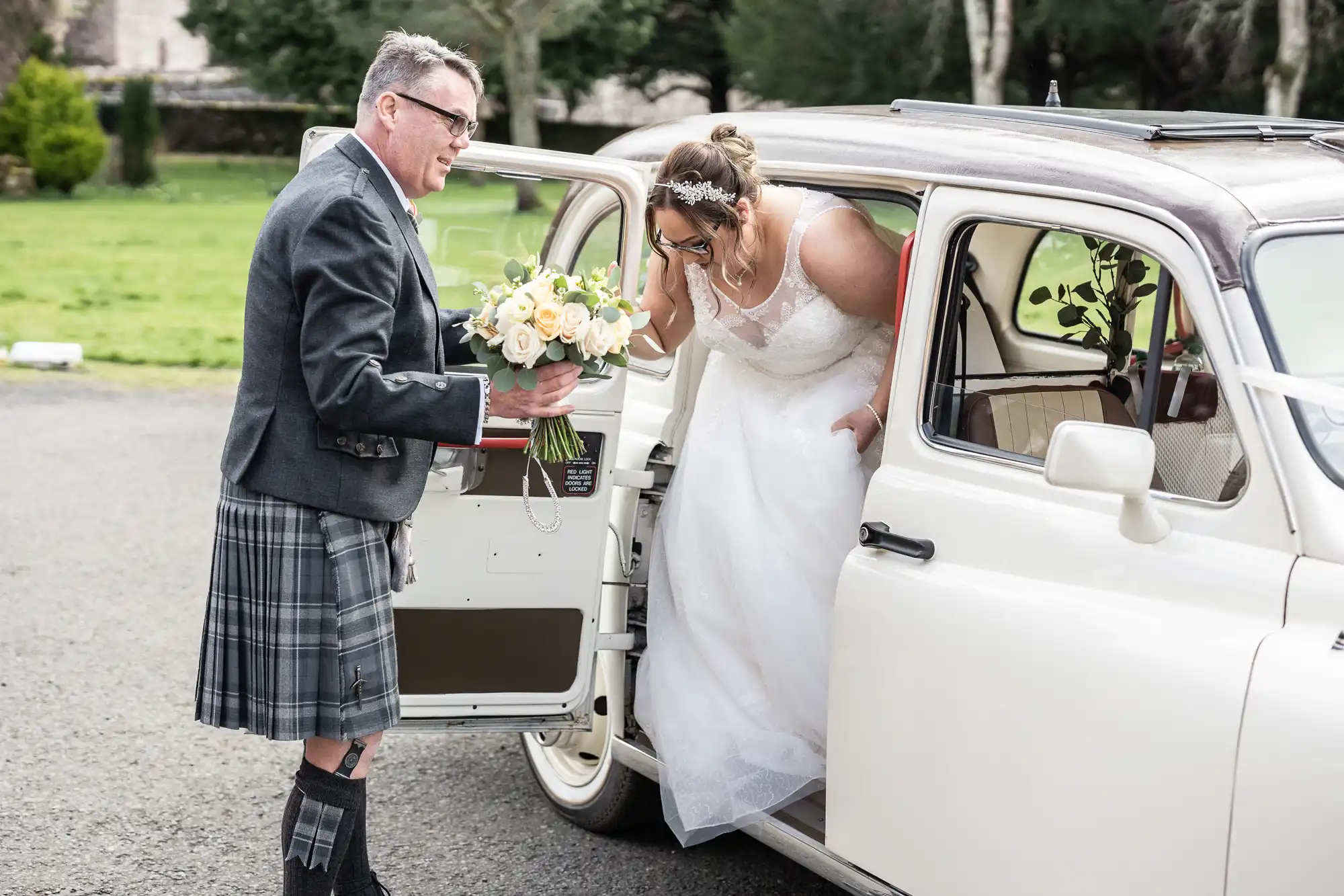 A man in a kilt holds a bouquet and helps a woman in a white wedding dress and tiara out of a vintage white car, parked on a paved area with grass and trees in the background. The woman leans forward, smiling as she leaves the car.