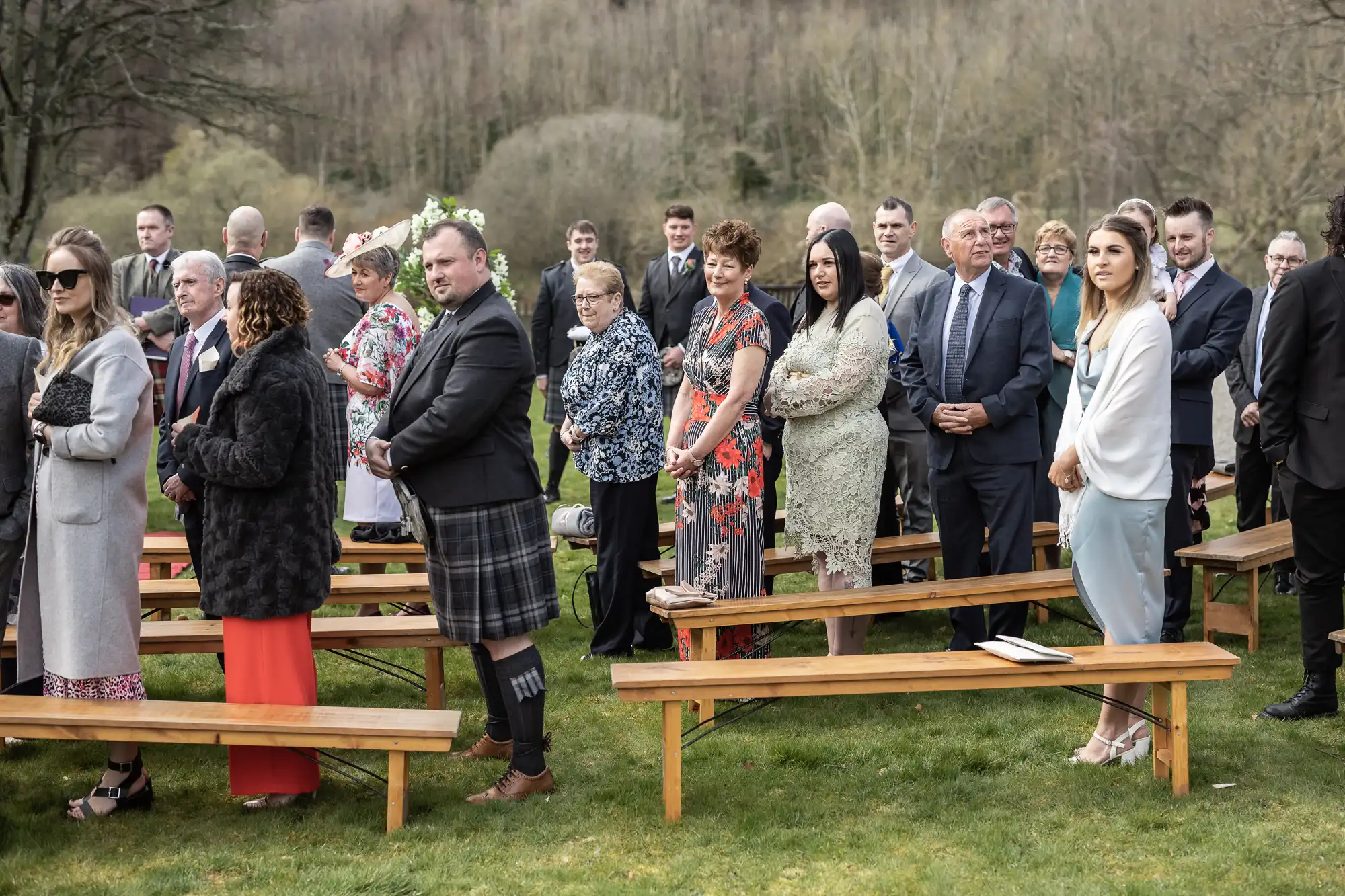 A group of well-dressed people, including men in suits and women in dresses and coats, stand outdoors on a lawn beside wooden benches, attending a formal event. Bare trees and hills are visible in the background, creating a natural setting.