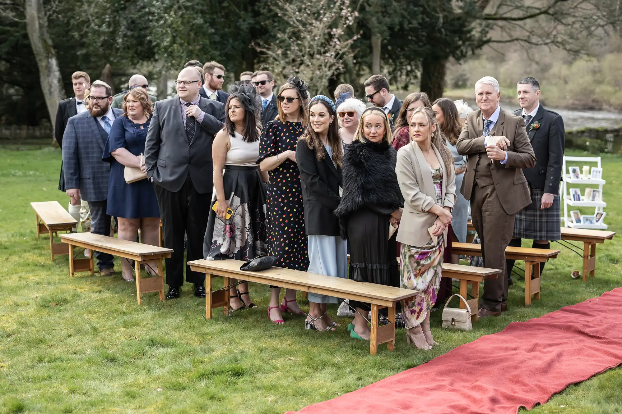 A group of formally dressed wedding guests stand and chat on grass beside wooden benches, near a red carpet. Trees and a small river are in the background, and some people wear sunglasses and coats, suggesting cool weather.