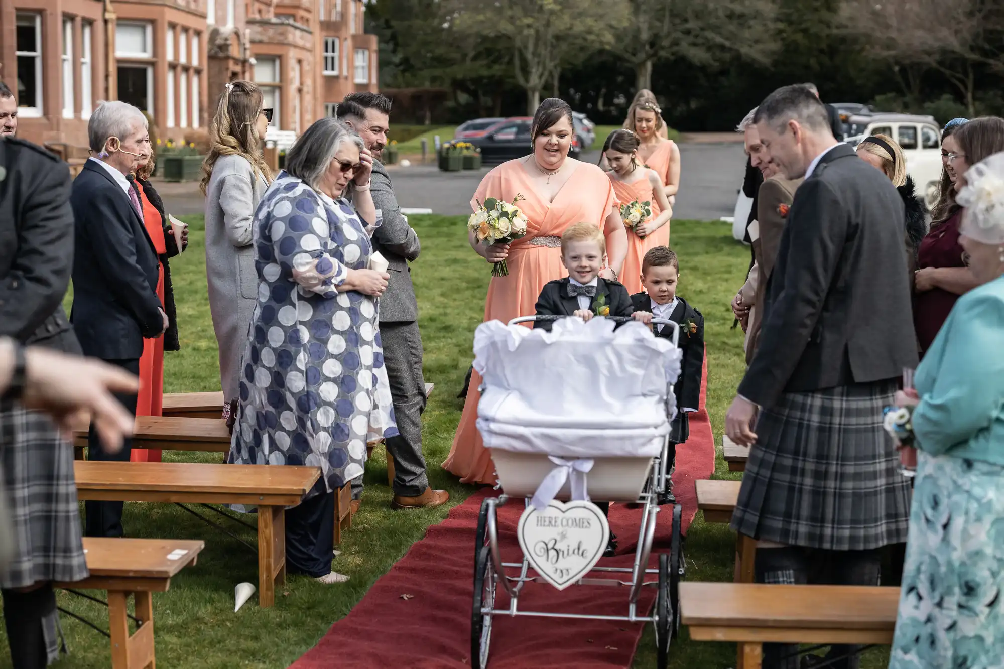 A wedding scene outdoors shows a smiling boy in a suit pushing a pram with a “Here Comes the Bride” sign, followed by another boy. Guests in formal wear line a red carpet, some watching and smiling. Bridesmaids in peach dresses stand in the background, holding bouquets.