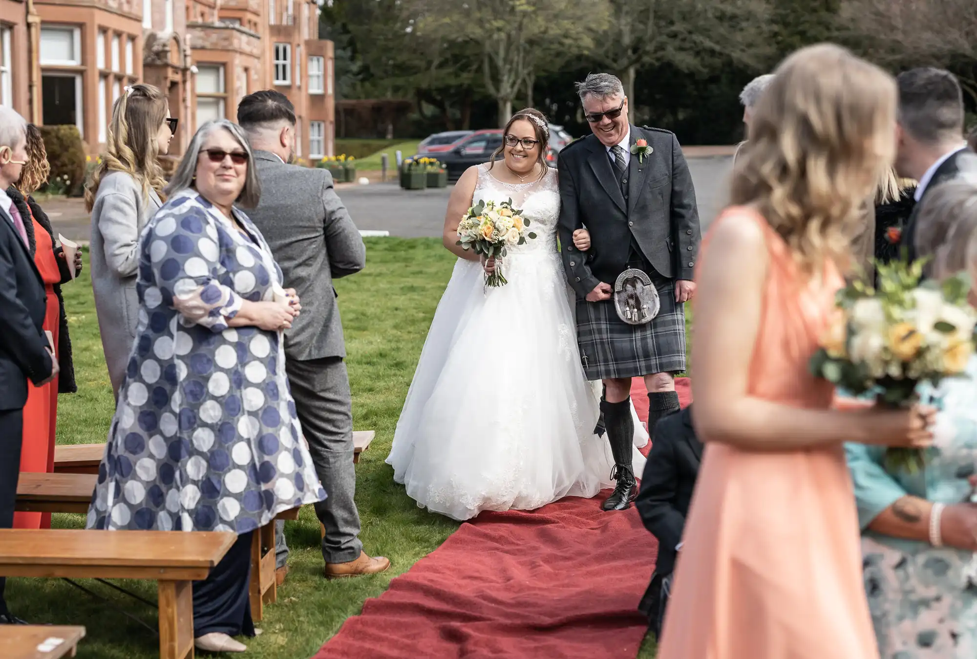 A bride in a white dress and a smiling man in a kilt walk arm-in-arm down a red aisle outdoors, surrounded by guests in colourful attire, some seated and some standing, with a brick building and trees in the background.