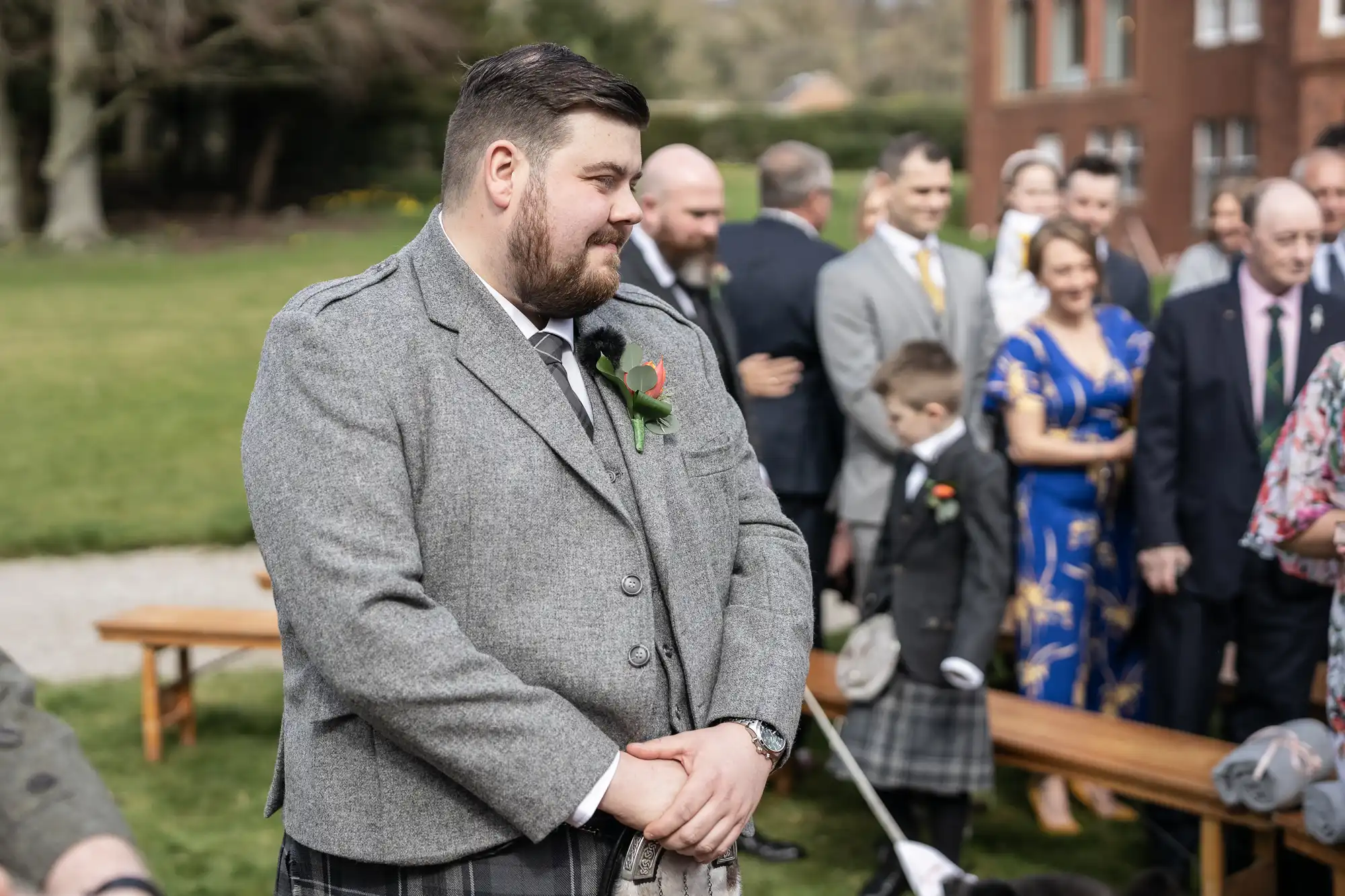 A man in a grey kilt suit stands outdoors, hands clasped, looking thoughtful. Behind him, people dressed formally, including a child in a kilt, are seated and standing on benches. Green grass, trees, and a building are visible in the background.