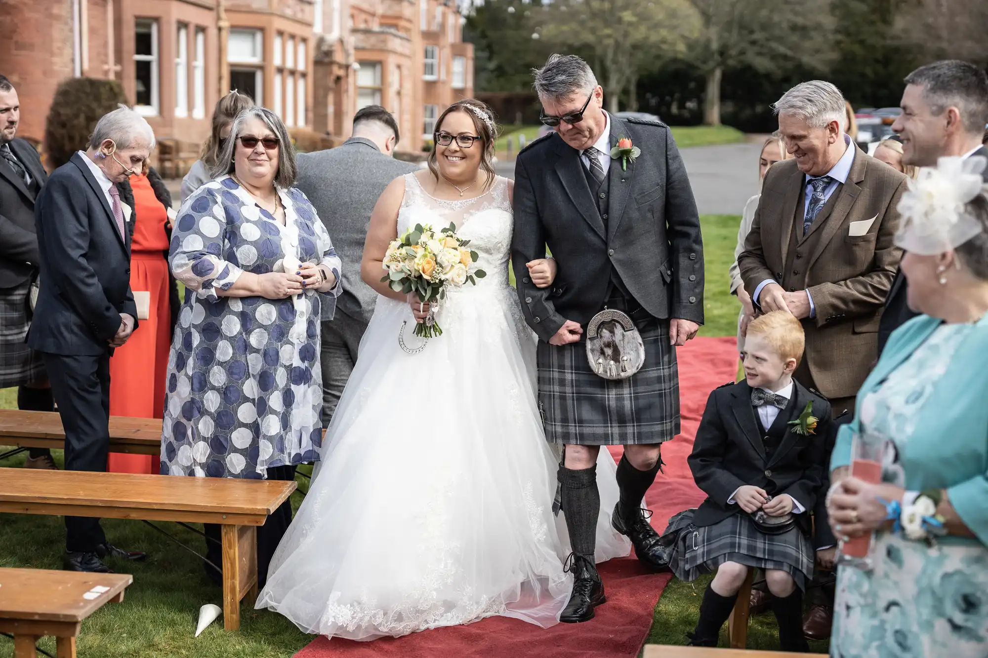 A bride in a white dress and glasses smiles as she walks down a red carpet outside, arm-in-arm with a man in a kilt. Guests, some in kilts and dresses, watch happily. The setting is grassy beside a large red-brick building under a cloudy sky.