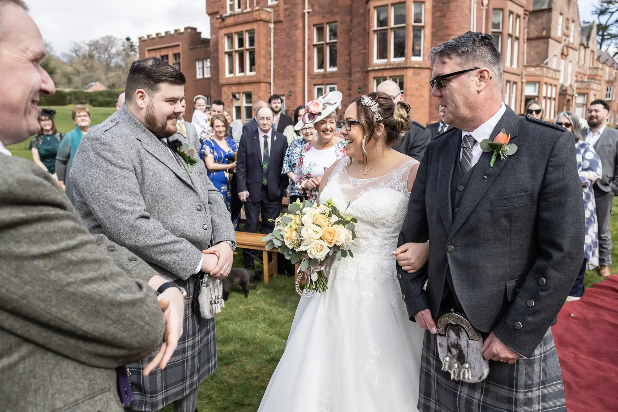 A bride in a white dress holding a bouquet walks arm-in-arm with an older man in a grey jacket and kilt, approaching a groom in a grey suit and kilt. Guests, smiling and dressed formally, watch outside a red brick building on a grassy lawn.