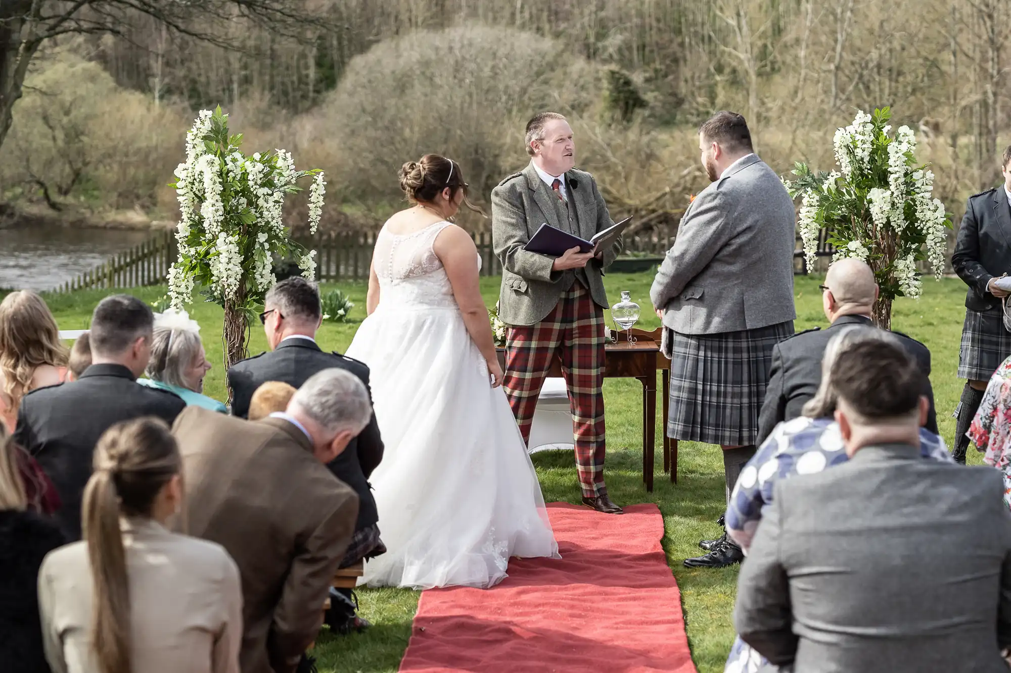 A bride and groom stand facing each other outdoors on a red carpet, with a male officiant between them. Guests are seated on either side. The scene features greenery, white floral arrangements, and trees in the background near a river.