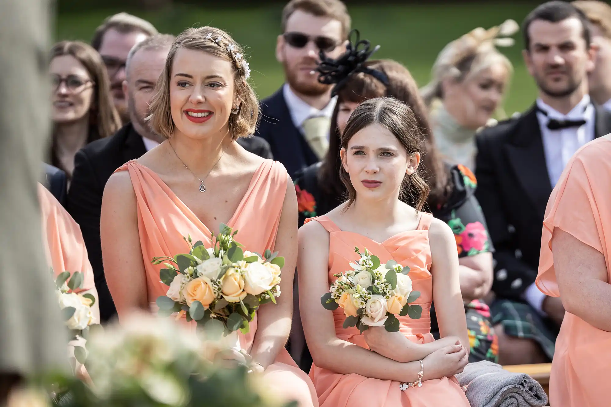 Two bridesmaids in peach dresses, holding flower bouquets, sit among other wedding guests outdoors. The woman on the left is smiling brightly; the younger girl beside her has a calmer expression. Guests in formal attire are seated in the background.