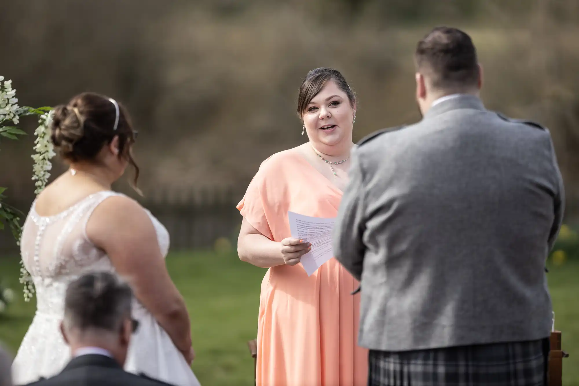 A woman in a peach dress reads from a piece of paper at an outdoor wedding ceremony, facing a bride in white and a groom in a grey suit. Another guest sits in the foreground. Greenery and blurred trees are in the background.