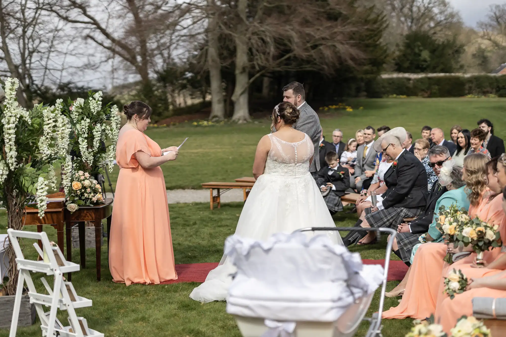 An outdoor wedding ceremony with a bride in a white dress and a groom standing before an officiant in a peach dress. Guests sit on wooden benches, and bridesmaids in matching dresses line the aisle. A white pram is in the foreground; trees and flowers fill the background.