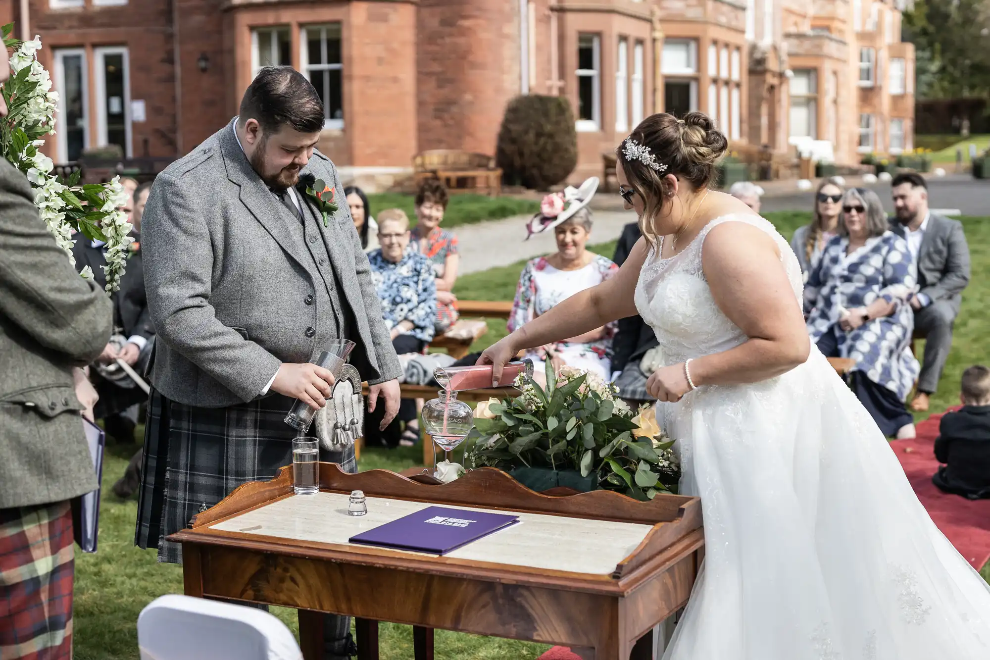 A bride in a white gown and a groom in a tartan kilt stand at a wooden table outdoors, pouring sand into a glass jar in a unity ceremony. Guests are seated on benches, and a large brick building is visible in the background. The scene is festive and joyful.