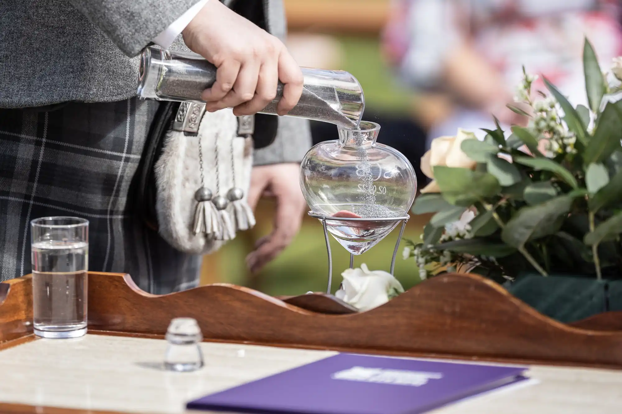 A person in a checked kilt pours water from a glass carafe into a round glass vessel on a wooden tray. A glass of water, a small empty glass, flowers, and a purple booklet are also on the tray. The setting is outdoors.