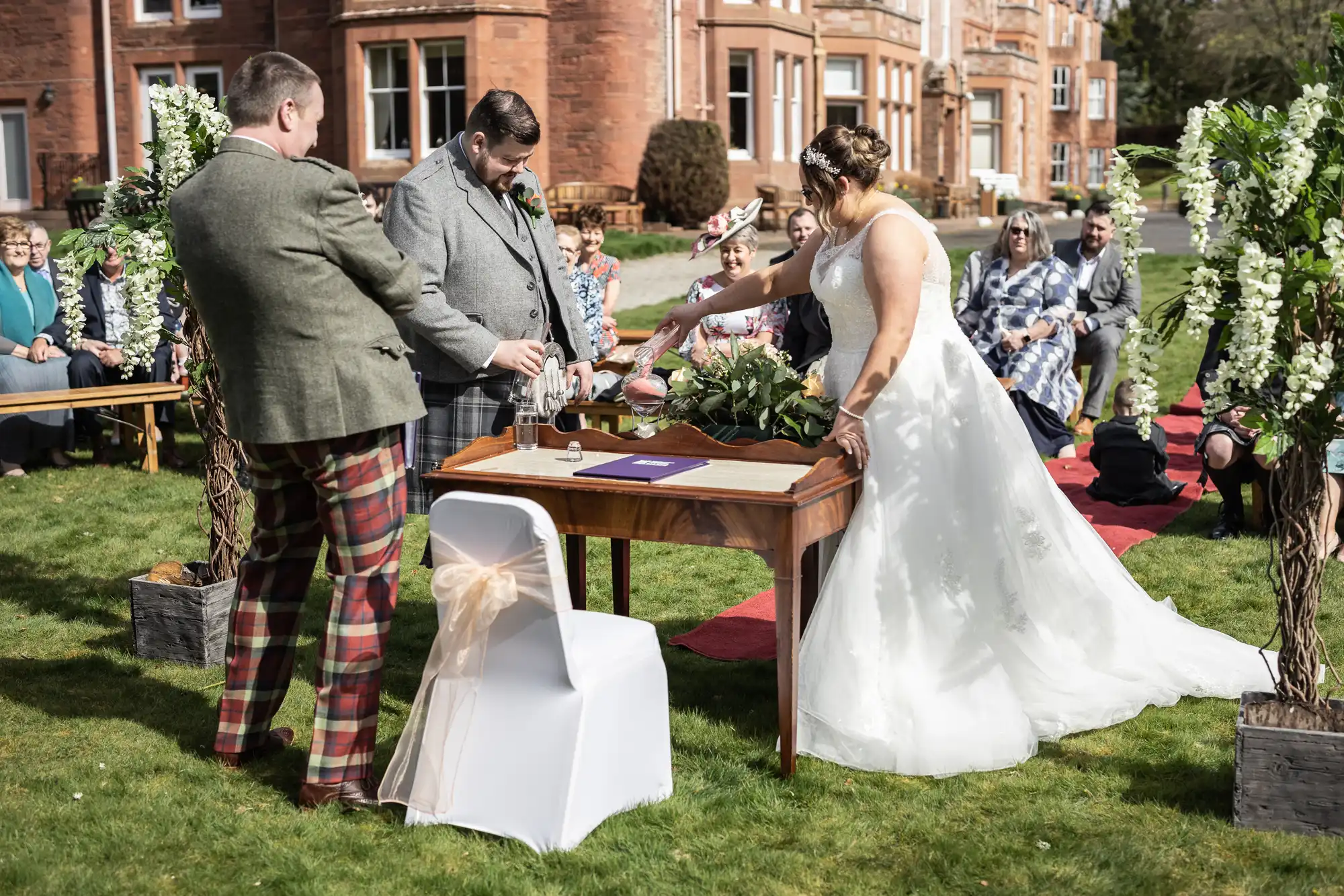 A bride in a white dress and two men in suits stand at a table outdoors, signing papers during a wedding ceremony. Guests sit on benches on a lawn, with flower arrangements and a large stone building in the background. The scene is bright and festive.