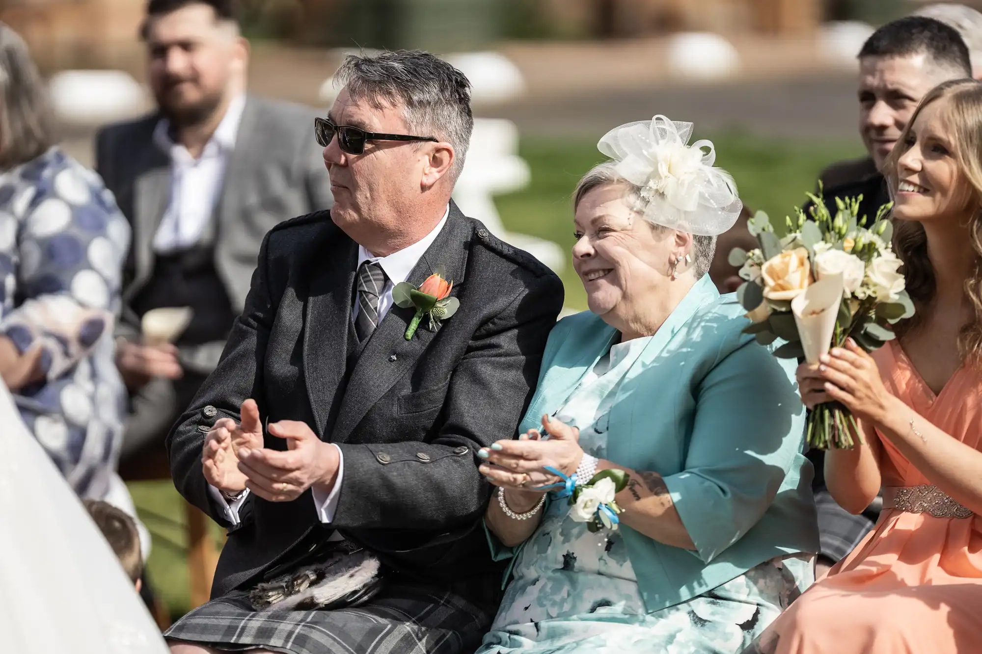 A smiling older couple sit outdoors at a wedding, applauding. The man wears sunglasses, a dark jacket, and a kilt, while the woman in aqua attire with a floral fascinator holds a corsage. Next to them, a woman in peach holds a bouquet. Other guests are blurred behind them.