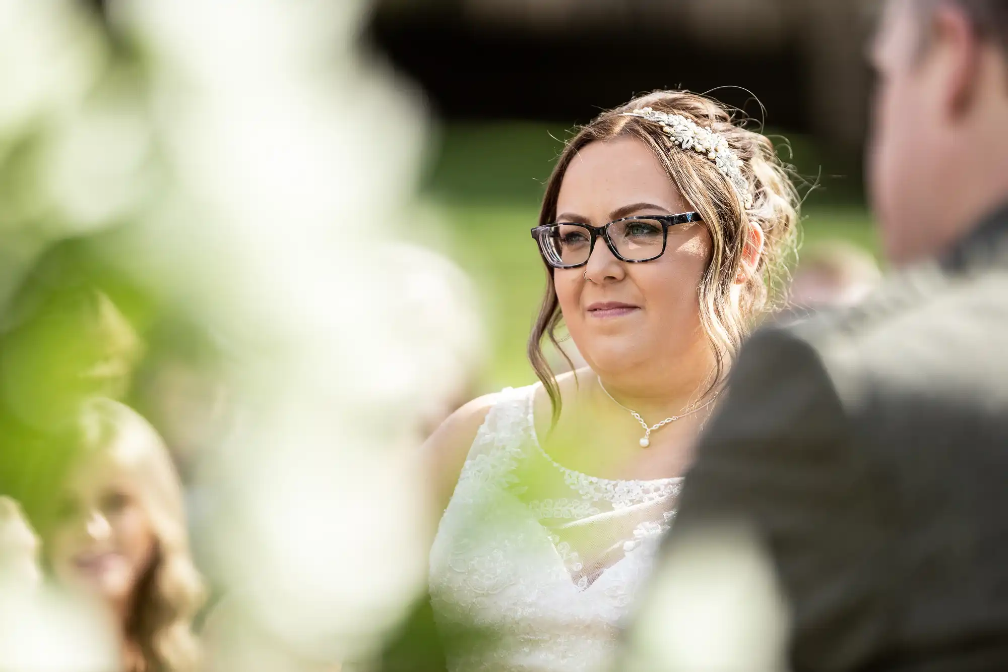 A bride with glasses, wavy hair, and a jewelled hairpiece looks forward during an outdoor wedding ceremony. She wears a white lace dress and a delicate necklace. Soft sunlight and green foliage create a bright, natural background.