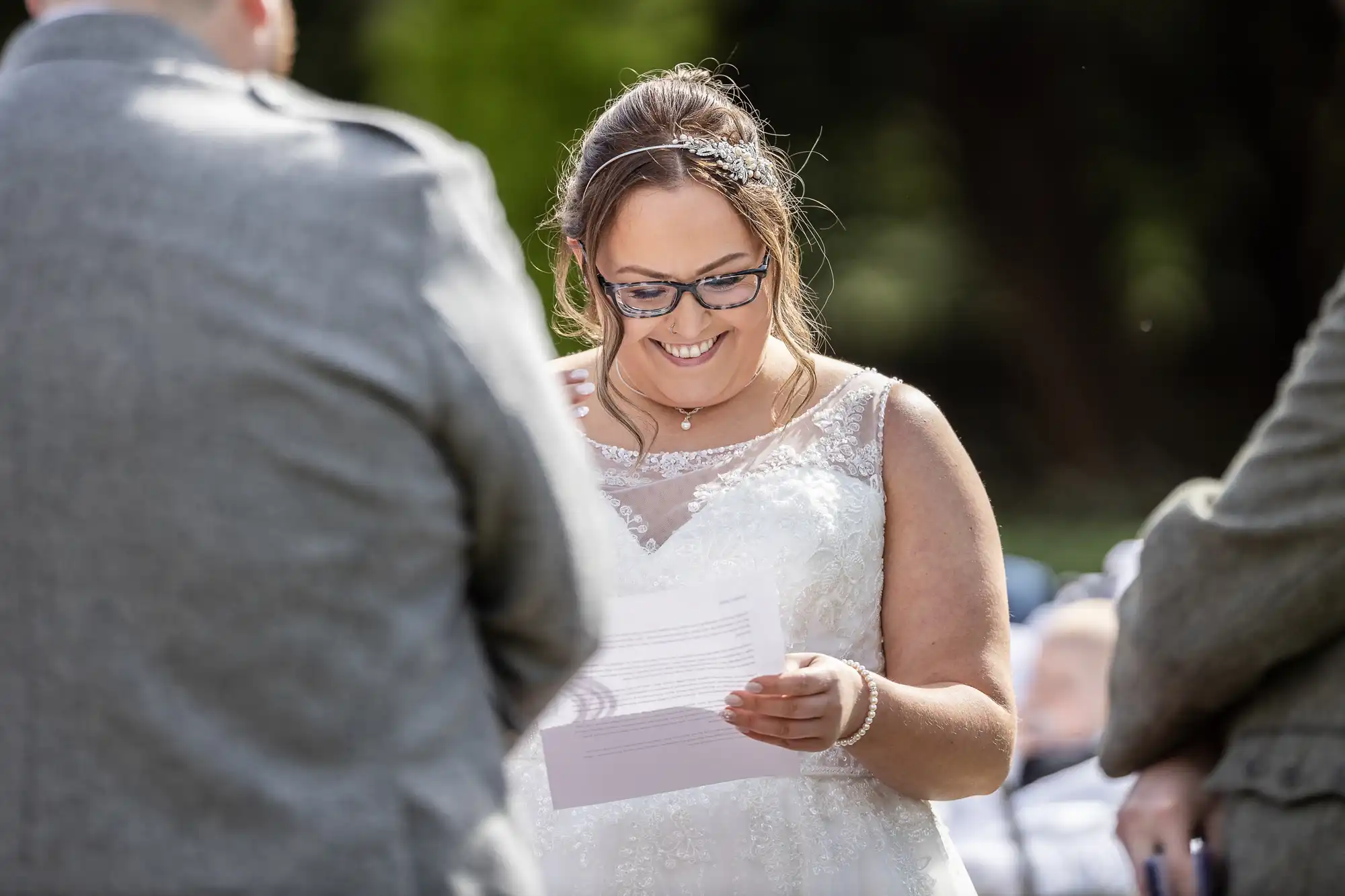 A smiling bride in a white lace wedding dress and glasses reads from a sheet of paper during an outdoor ceremony, flanked by two people in grey suits; blurred greenery is visible in the background.