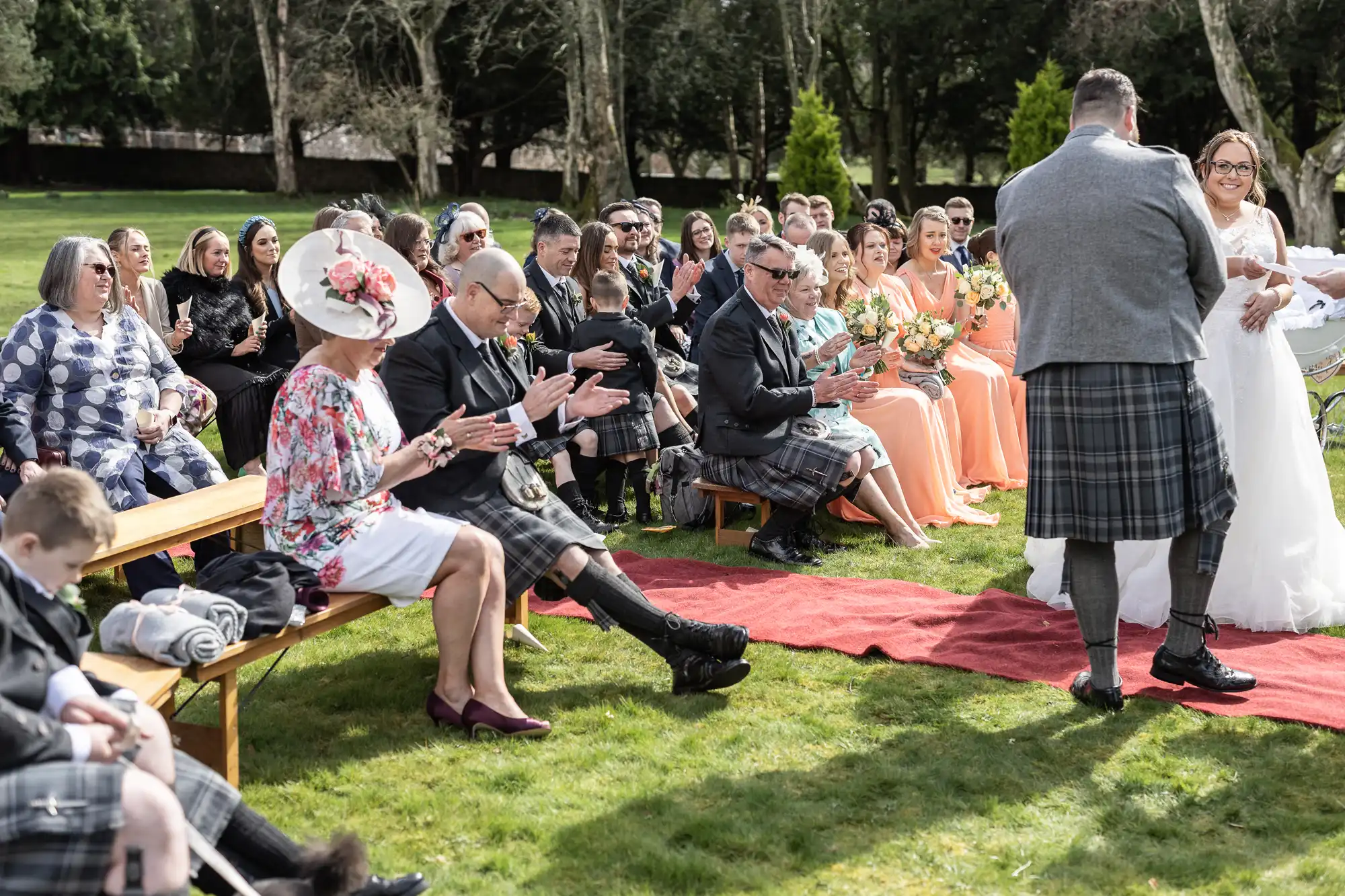 An outdoor wedding ceremony; guests sit on benches, many in kilts or formal wear, applauding as a smiling bride in a white dress holds hands with a groom in a grey jacket and kilt. Bridesmaids in peach dresses are seated at the front. Lush green lawn and trees surround them.