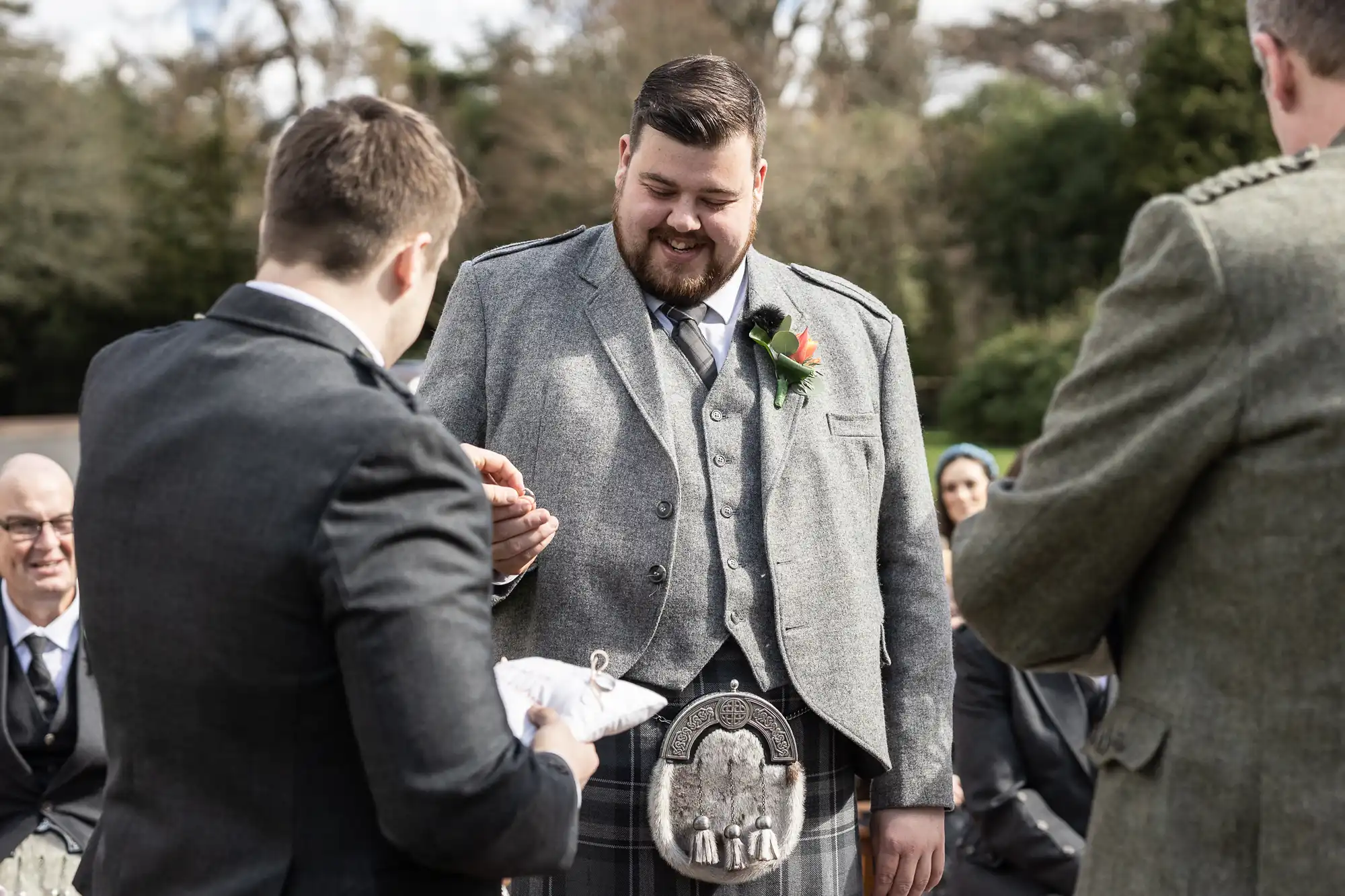 A smiling groom in a grey kilt suit places a ring on his partner's finger during an outdoor wedding ceremony. He wears a sporran and buttonhole. Guests in suits look on, seated, with trees and greenery blurred in the background.