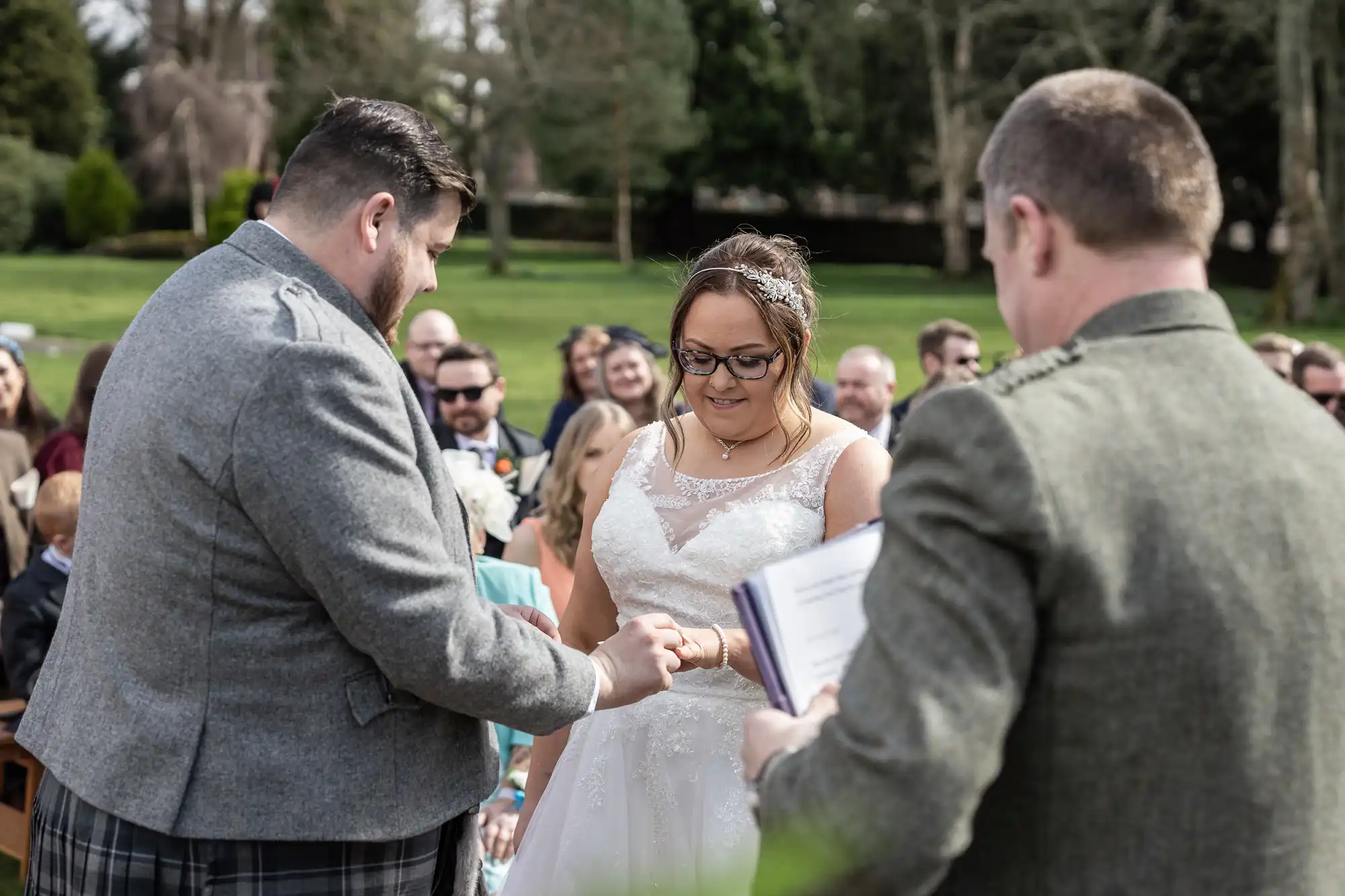 A groom places a ring on the bride’s finger during an outdoor wedding ceremony. The bride wears a white dress and glasses, smiling. Guests are seated in the background, and an officiant holds a book in the foreground. The scene is set in a green park.